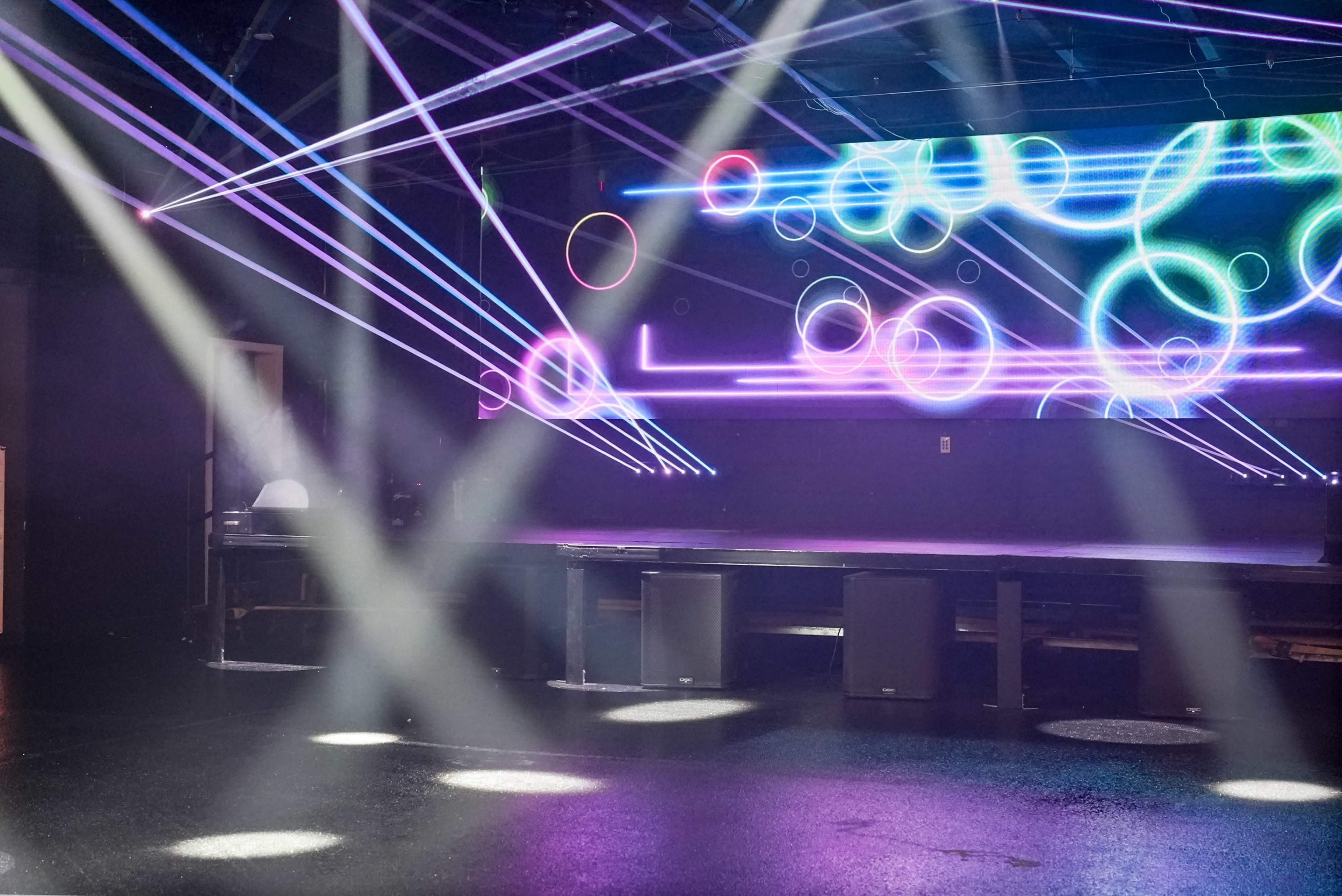 The image shows a dimly lit stage with colorful laser lights projecting geometric patterns against a backdrop.