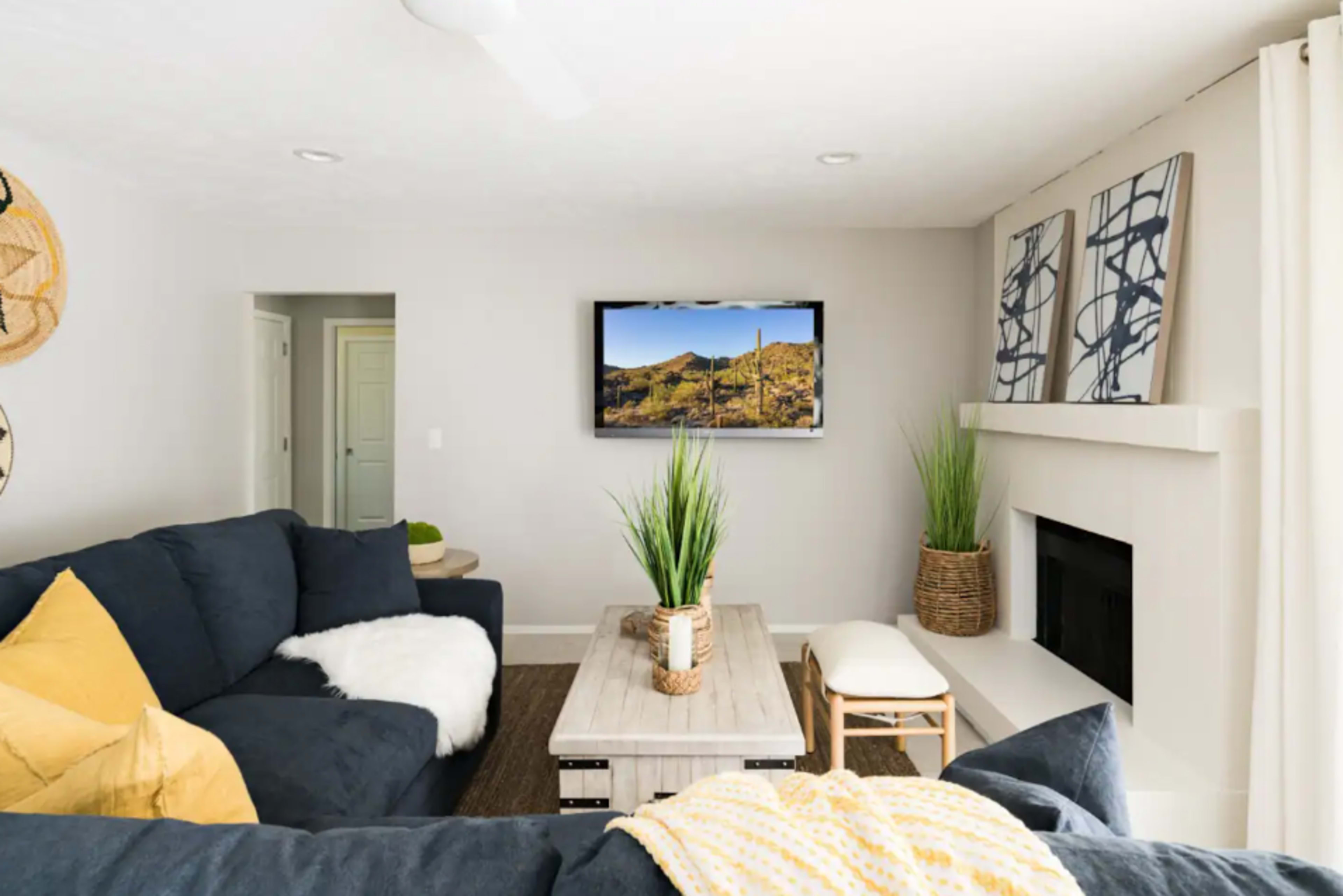 A modern living room features a dark sofa, a wooden coffee table, potted plants, and a wall-mounted television displaying a desert landscape.