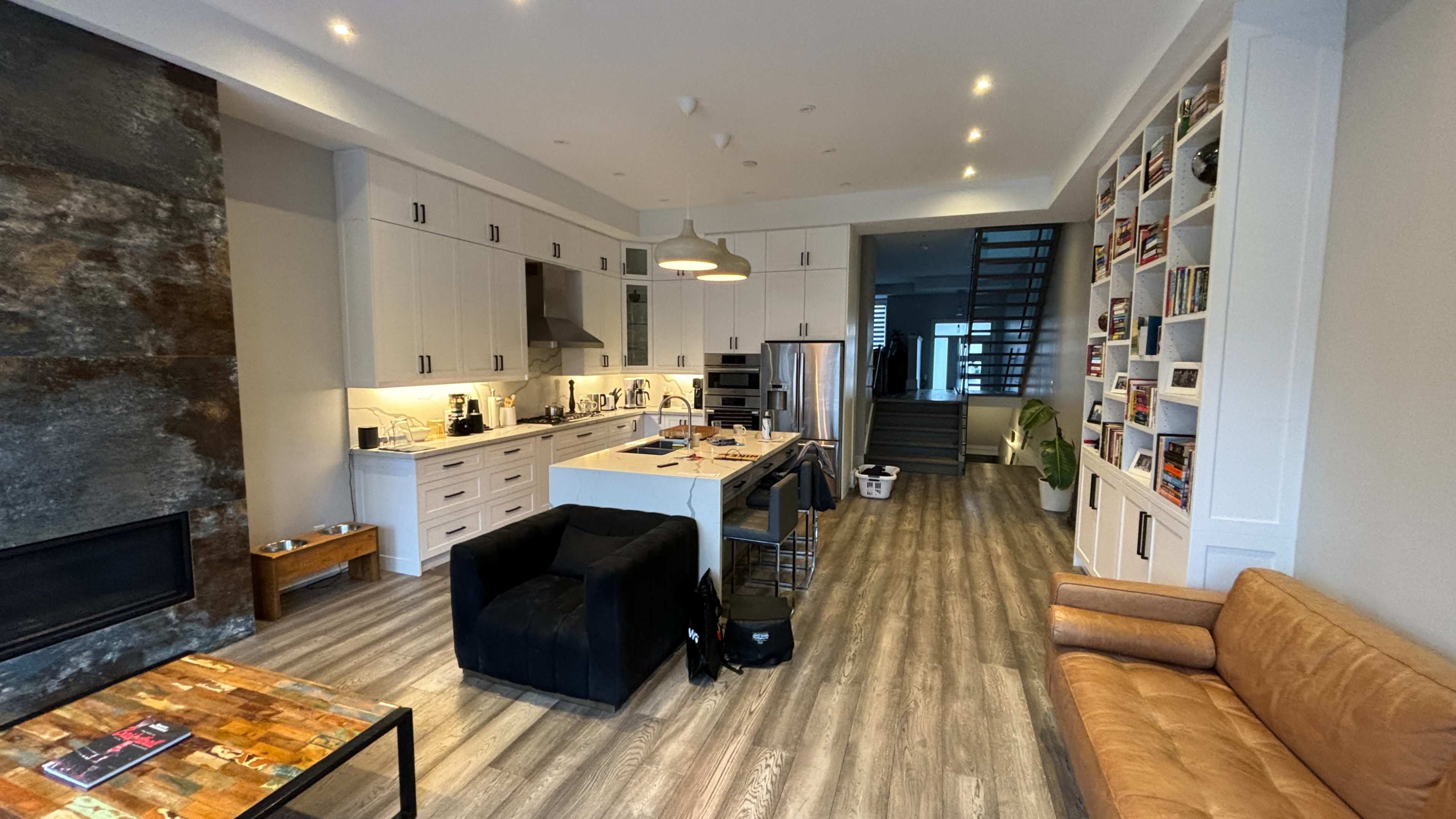 A modern kitchen and living room area with a large island, black chairs, and a staircase in the background.