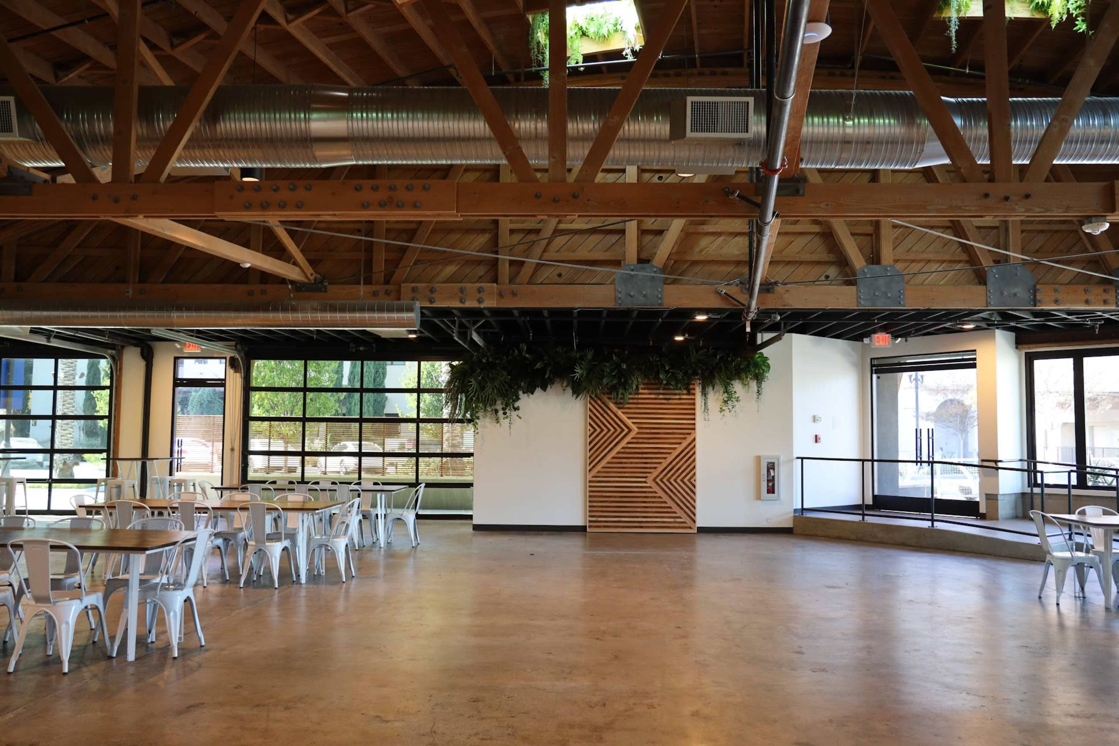 The image shows a spacious interior of a modern venue with wooden beams, large windows, and white tables and chairs arranged in an open area.