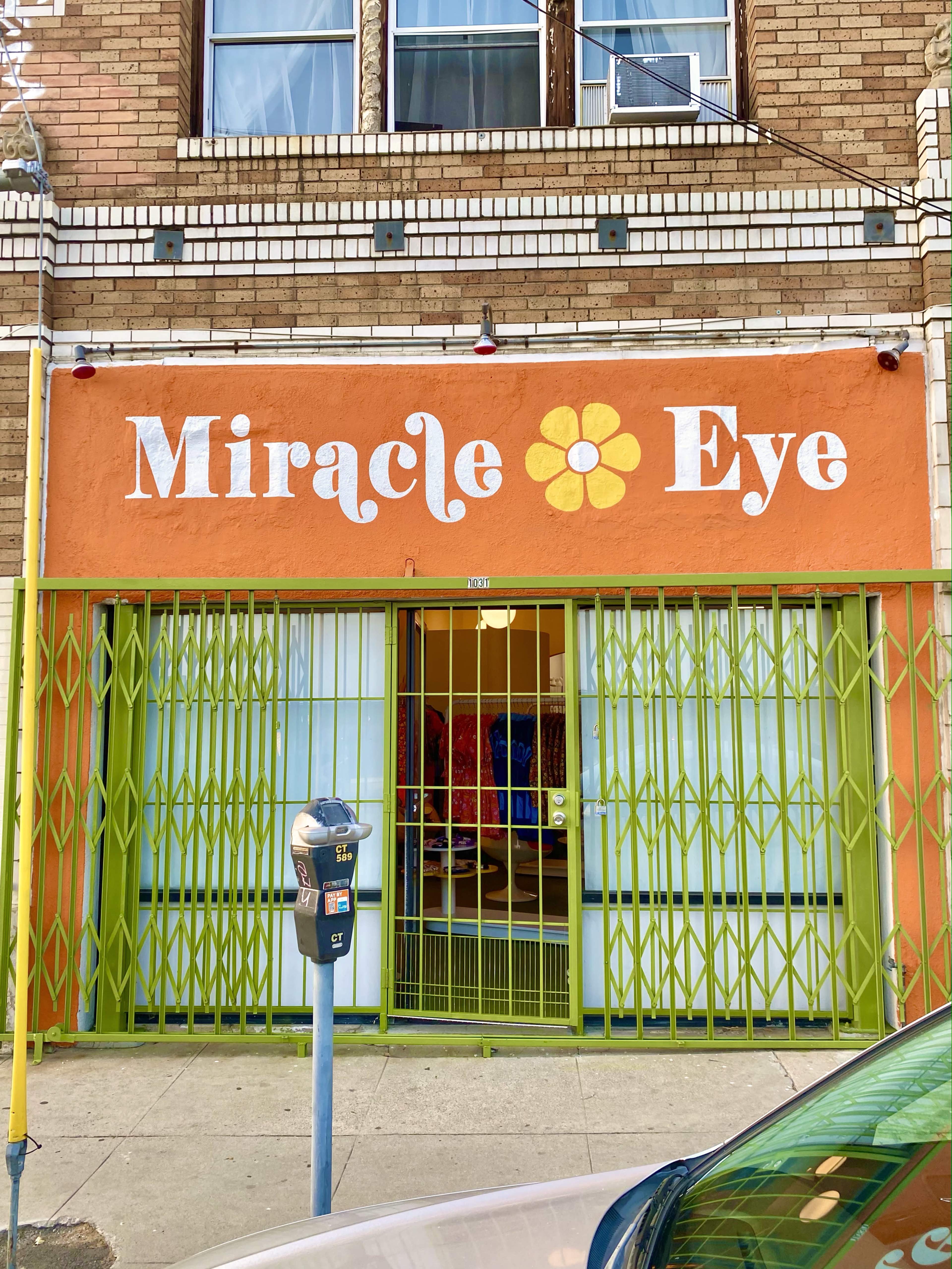 The storefront of "Miracle Eye" features a large orange sign with a flower icon, surrounded by green bars and a parking meter.