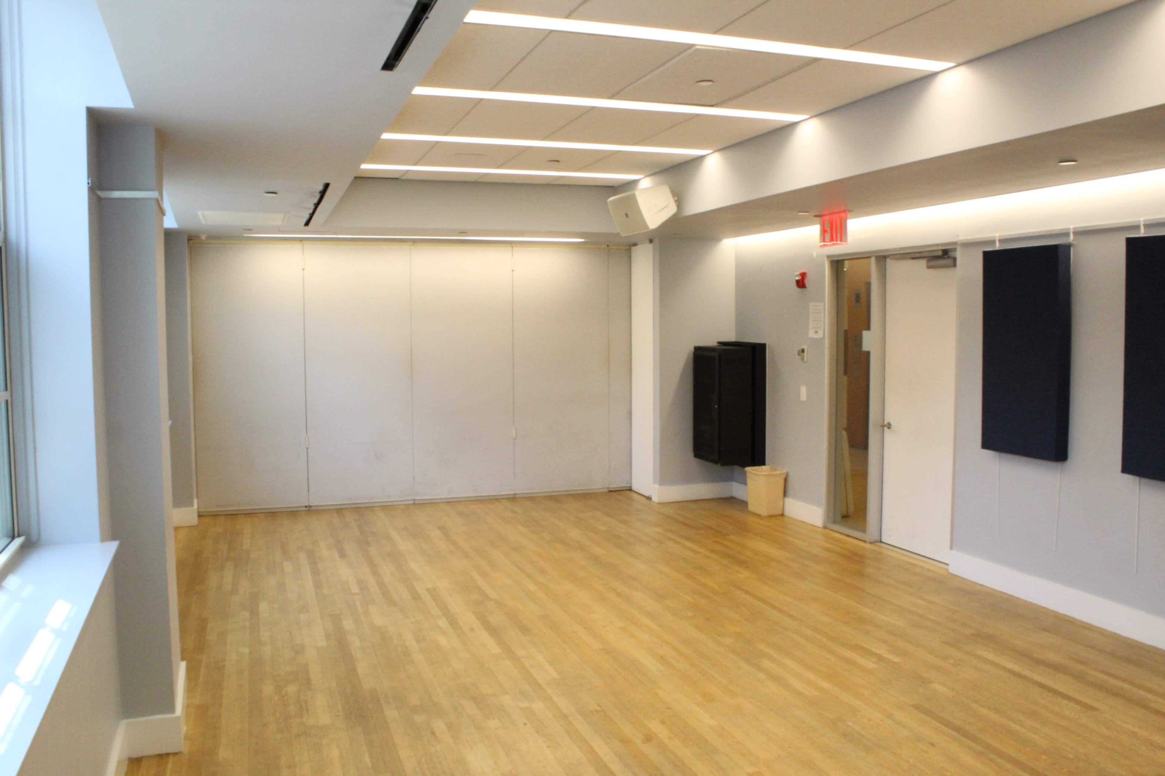 The image shows a spacious, well-lit room with wooden flooring and gray walls, featuring a projector and sound equipment mounted on the ceiling.