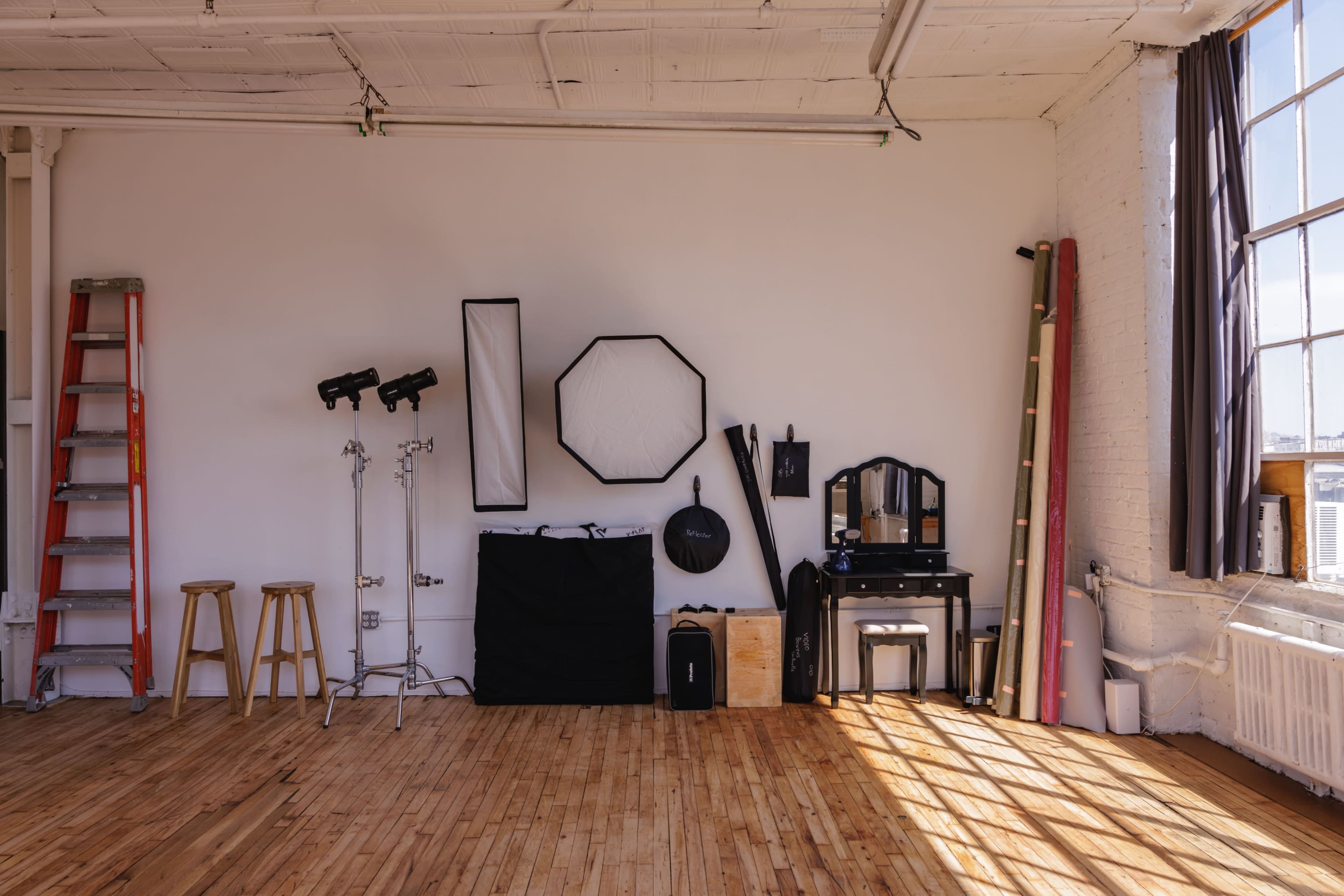 The image shows a photography studio with a wooden floor, featuring lighting equipment, backdrops, a large octagonal softbox, a mirror, and a ladder against a white wall.