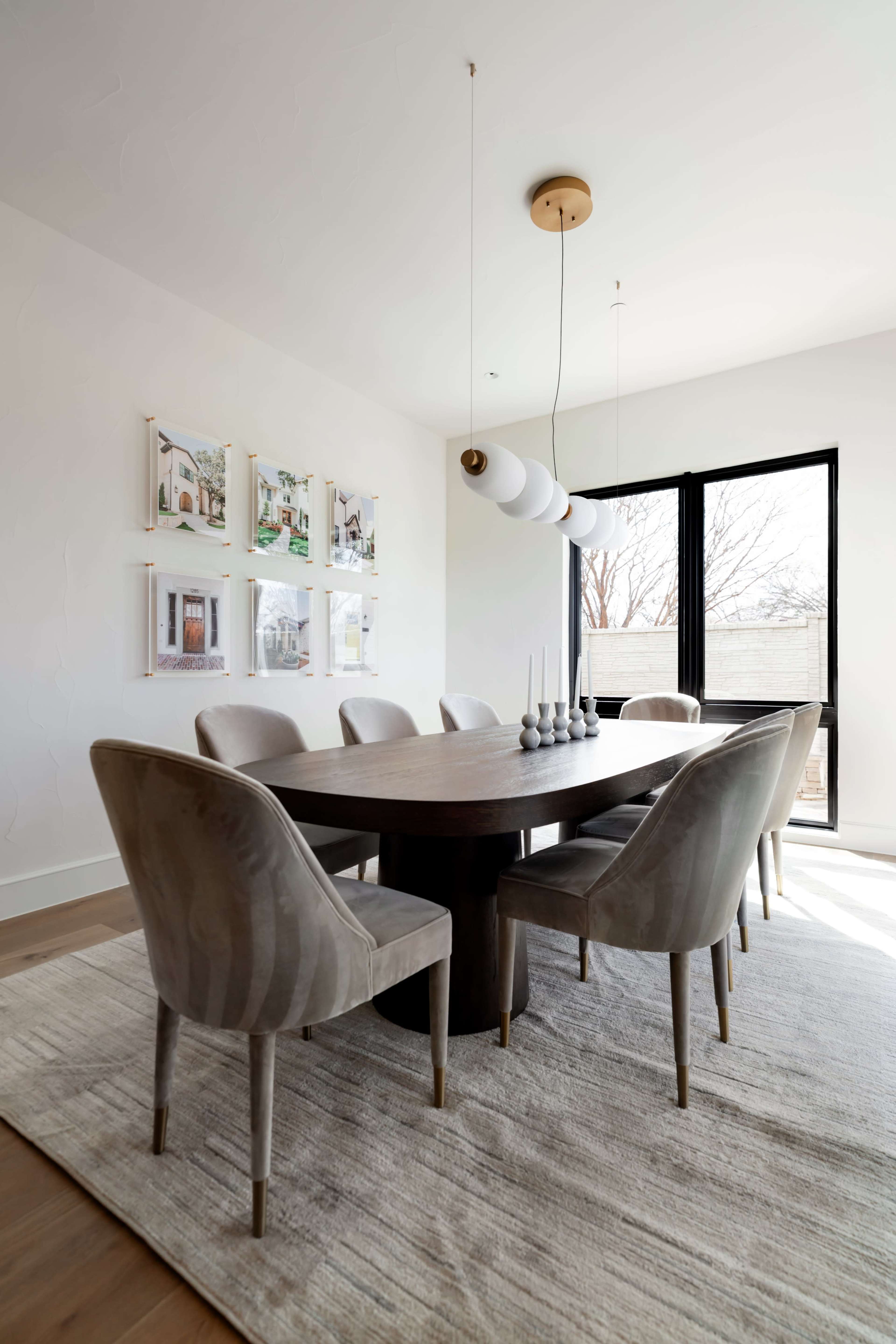 A modern dining room features a round wooden table surrounded by six upholstered chairs, with framed artwork on the wall and large windows letting in natural light.
