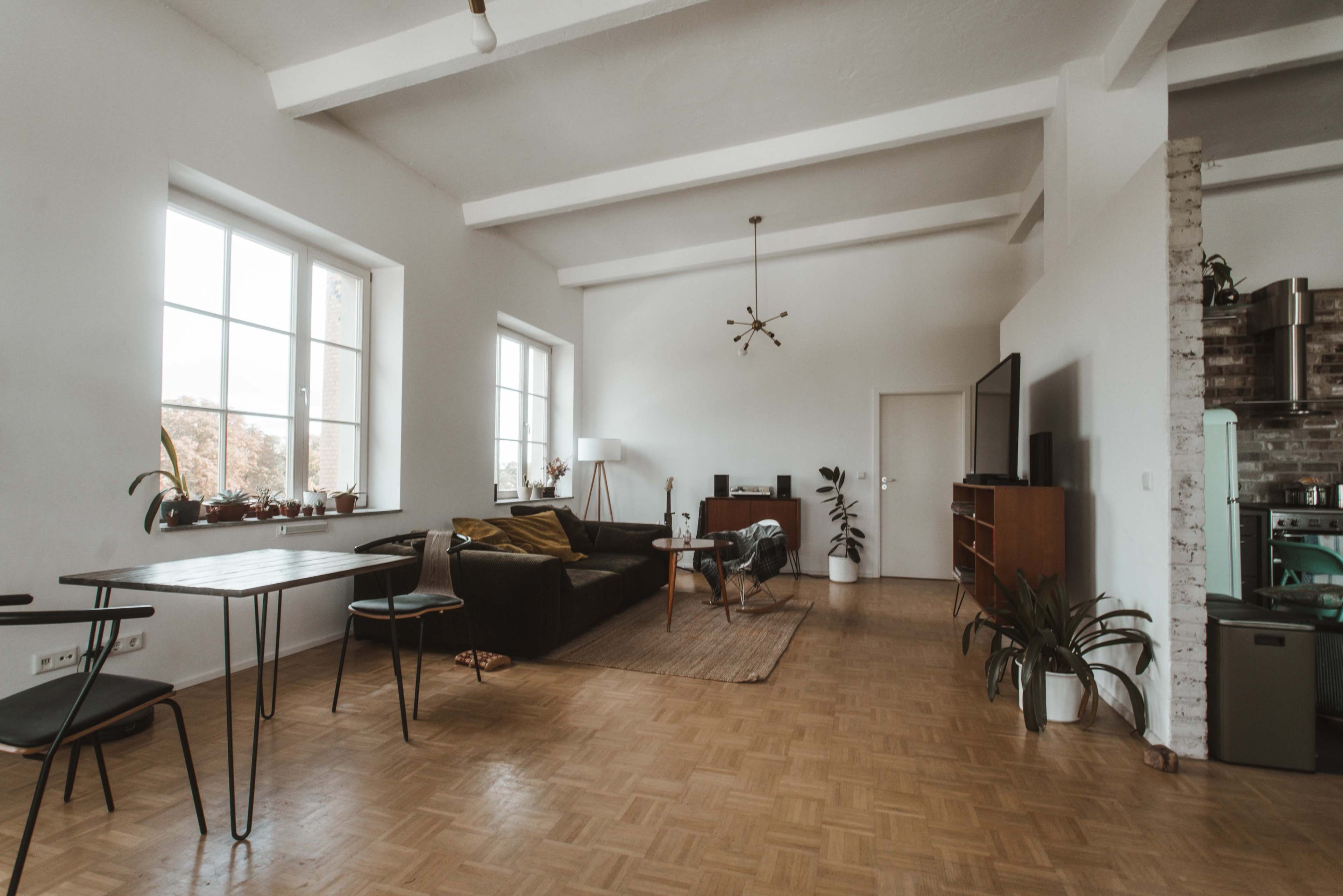 The image shows a spacious, modern living room featuring a sectional sofa, a dining table, and large windows allowing natural light to fill the space.