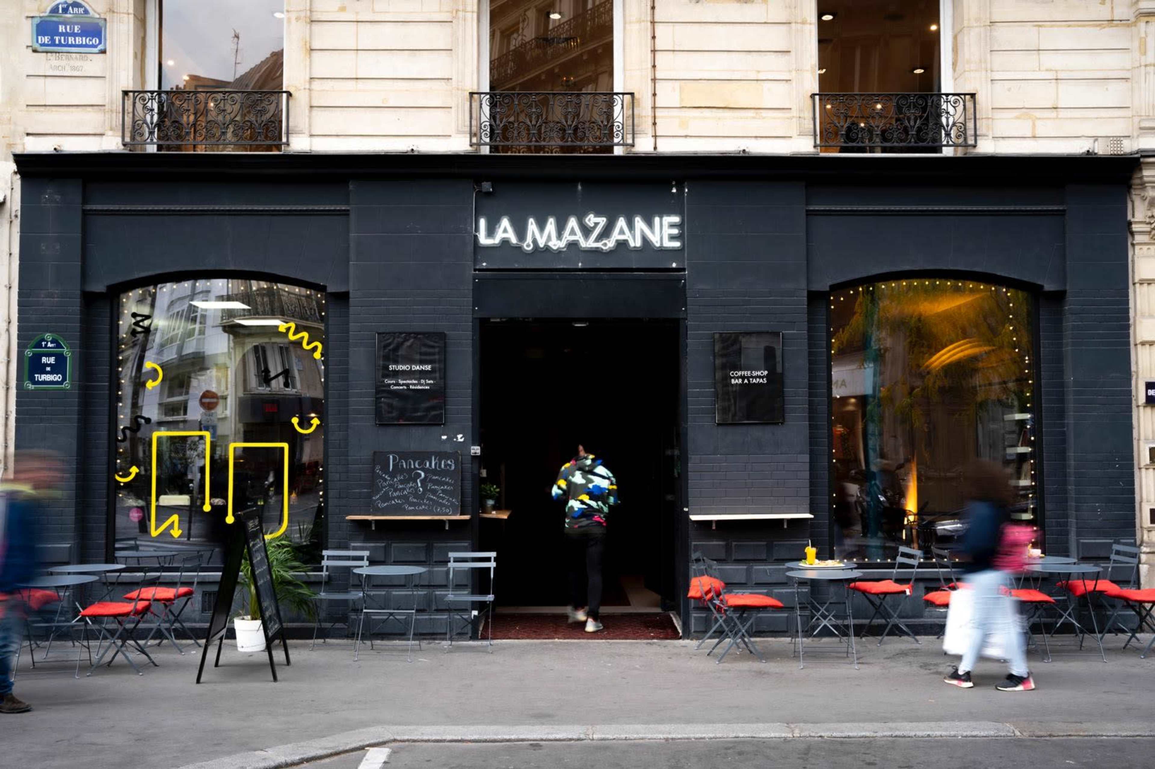 The image shows the entrance of a café named "La Mazane," featuring a black facade with large windows and outdoor seating arrangements.