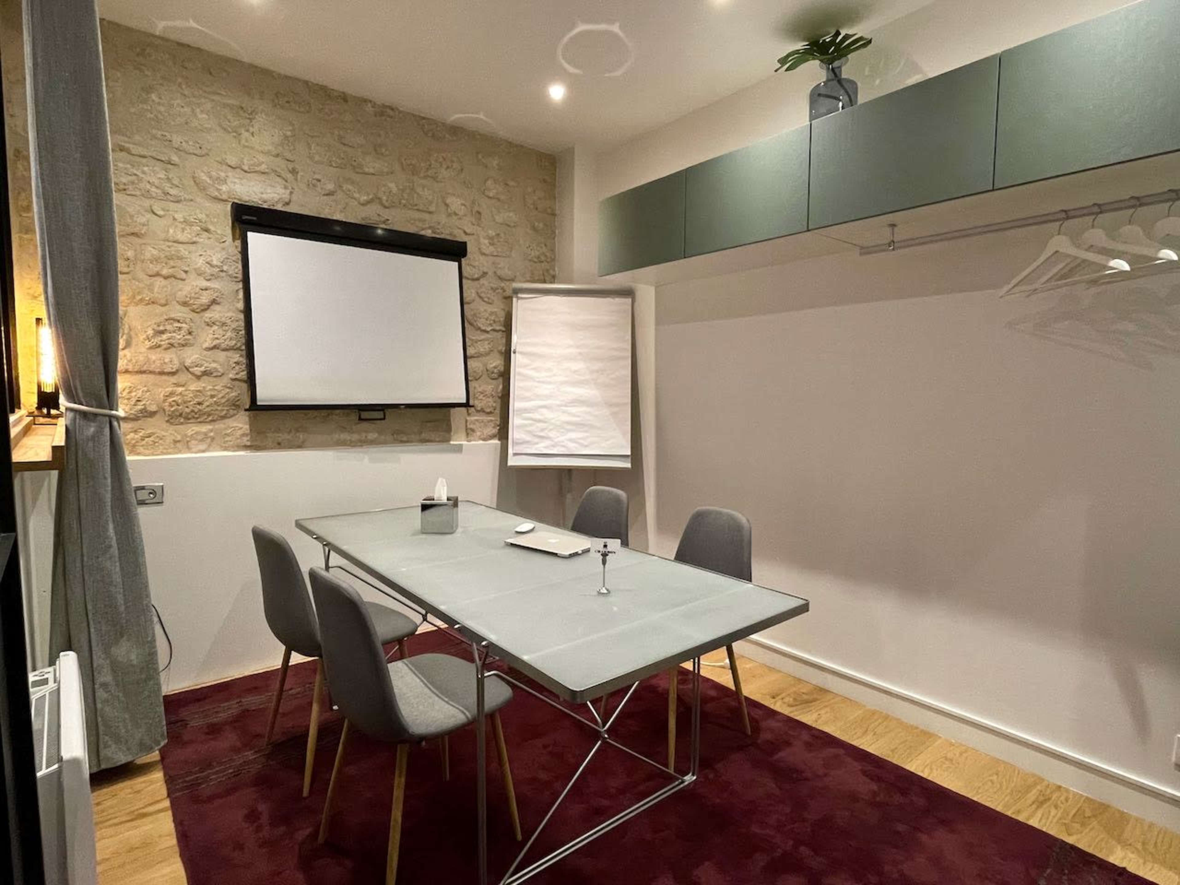 A meeting room features a rectangular glass table surrounded by six gray chairs, a projection screen, a flip chart, and a plant on a shelf.