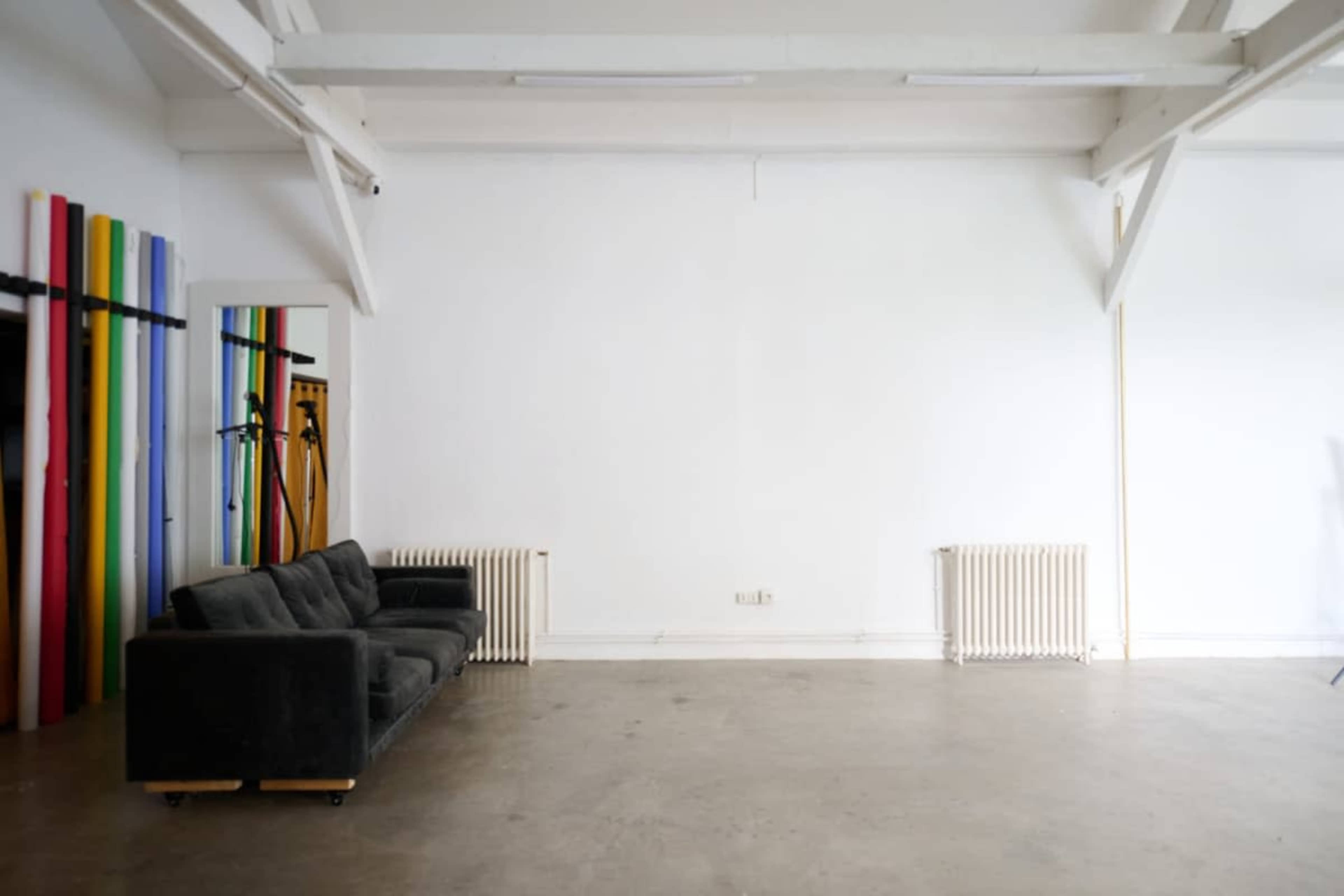 The image shows a spacious, empty room with a gray sofa, a radiator, and colorful vertical panels against one wall.