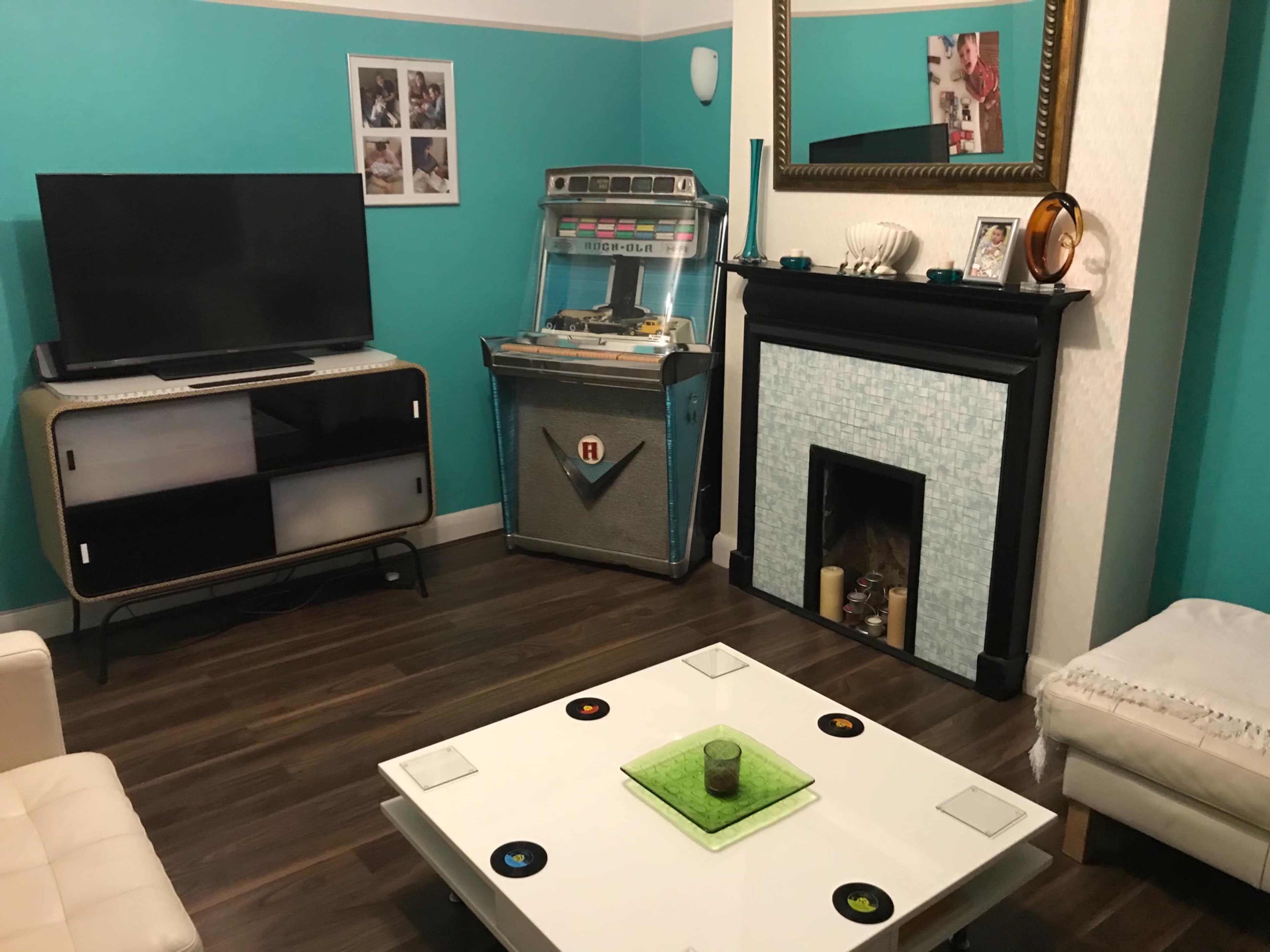 A living room with a television, a jukebox, and a coffee table, all set against teal walls and wooden flooring.
