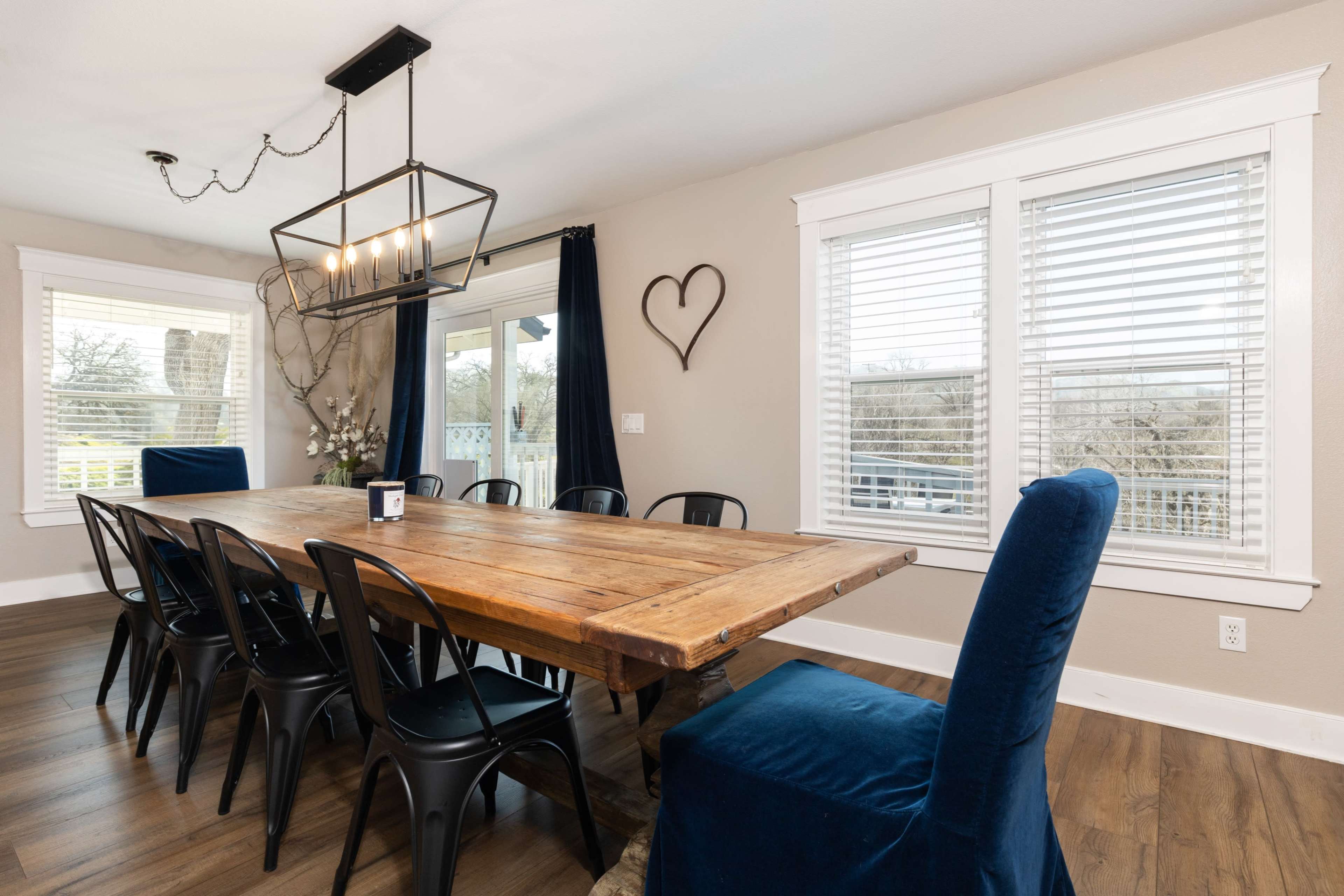 A large wooden dining table is set with black metal chairs and blue upholstered chairs, placed in a room with light walls, large windows, and a chandelier.