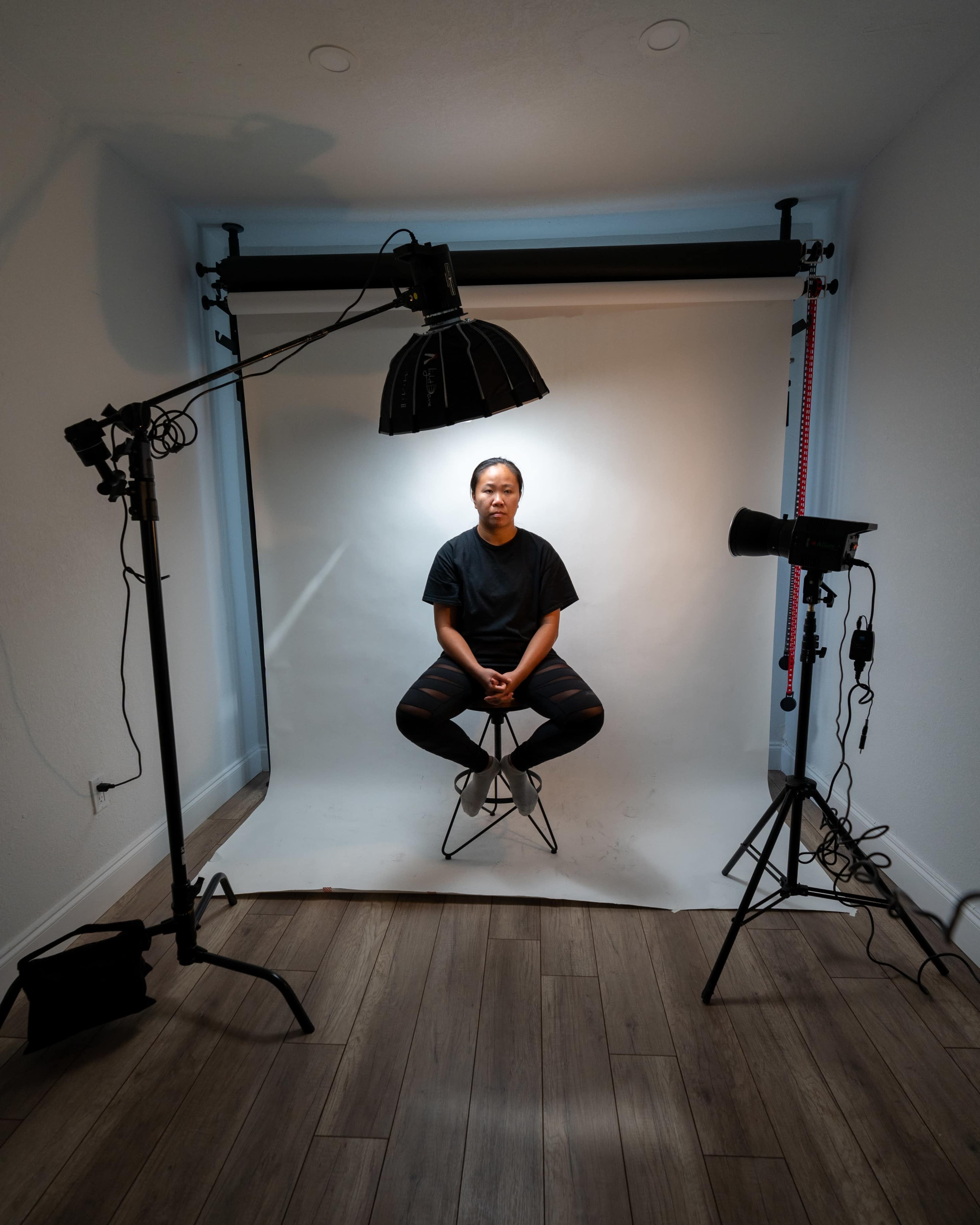 A person sits on a stool in a photography studio, surrounded by studio lights and a backdrop.