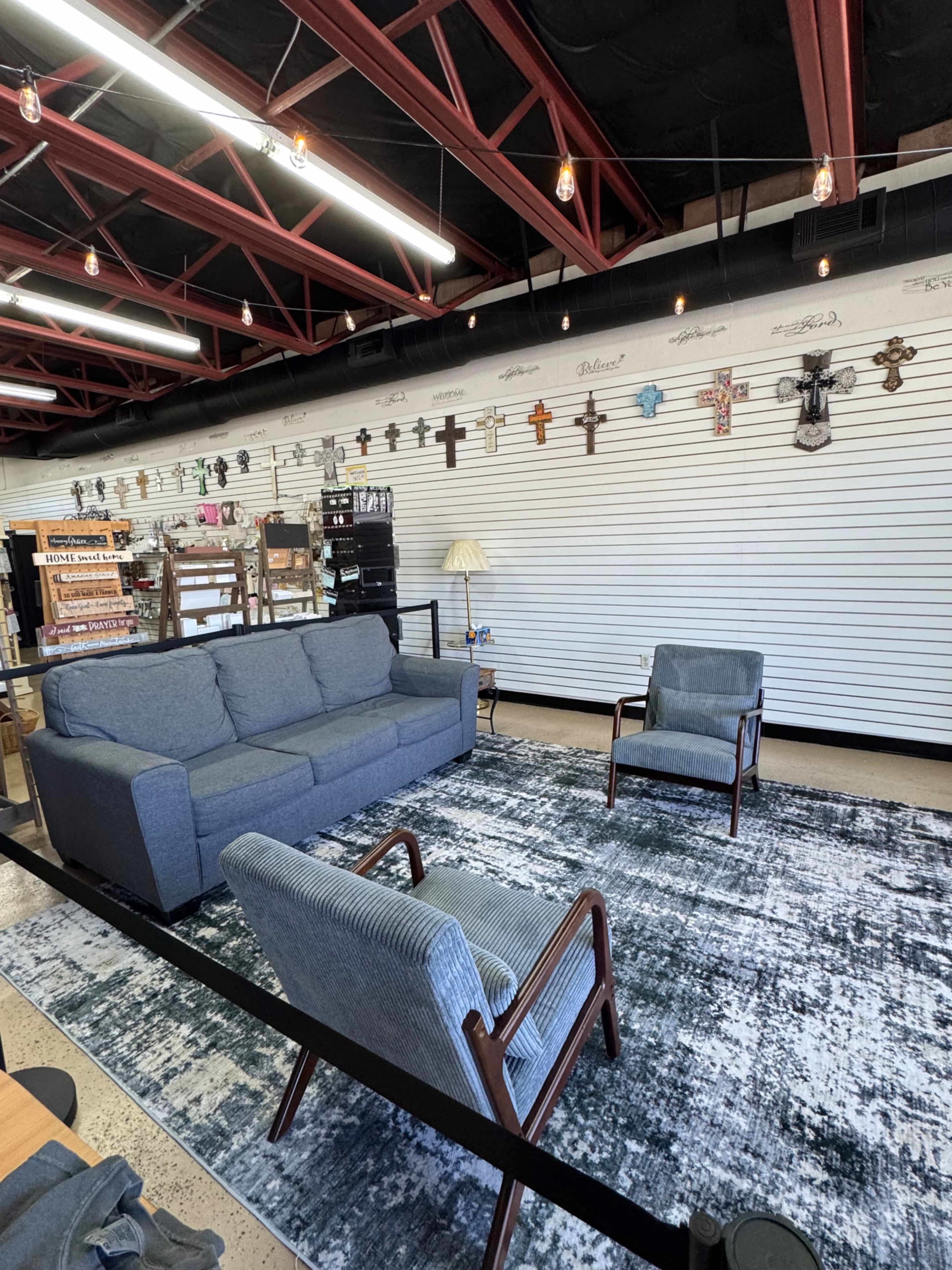 The image shows a furnished retail space featuring a gray sofa, two matching chairs, and a patterned area rug, with a wall displaying various decorative items.
