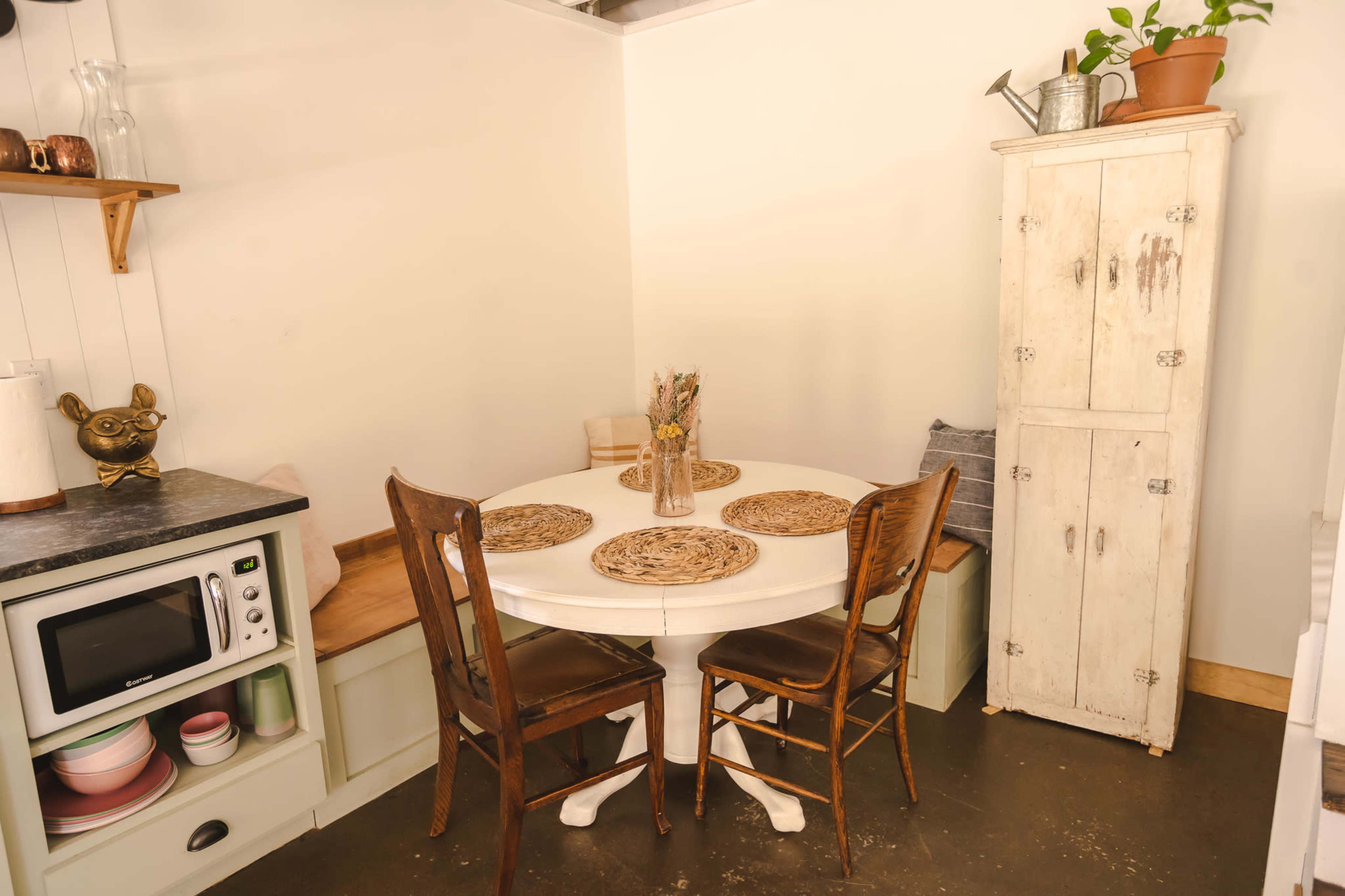 A small kitchen features a round dining table with four wooden chairs, surrounded by a cupboard, microwave, and decorative plants.