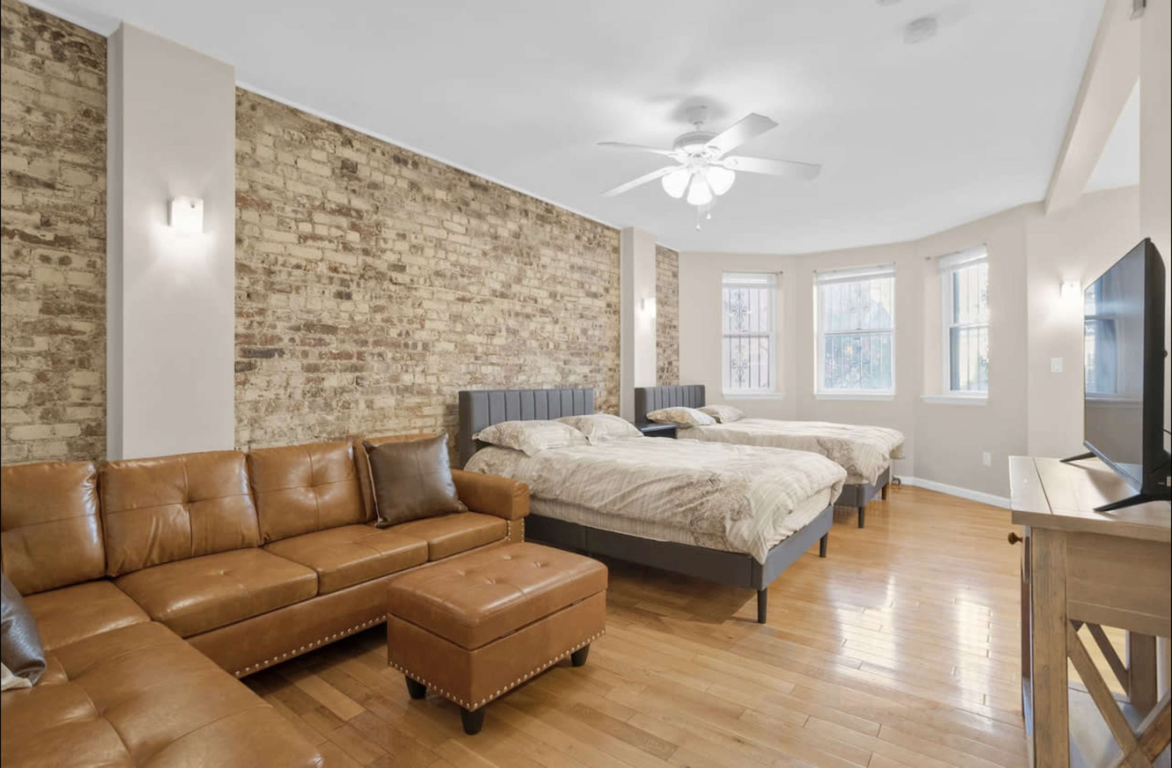 The image shows a spacious bedroom featuring two large beds, a brown leather sofa, and a wooden TV stand against a wall with exposed brick.
