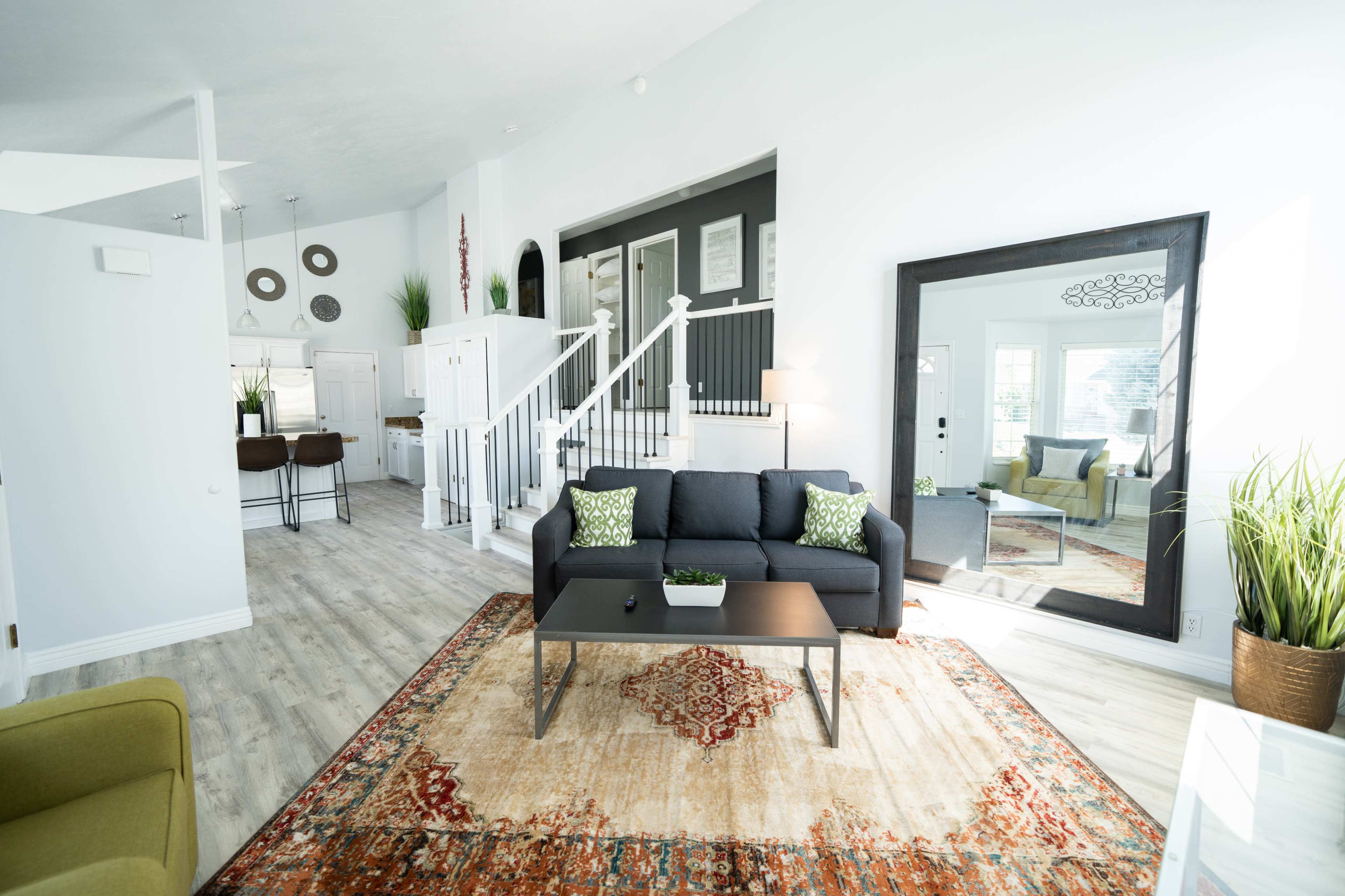 A living room features a gray sofa with green pillows, a coffee table, and a large mirror, complemented by a patterned area rug and houseplants.