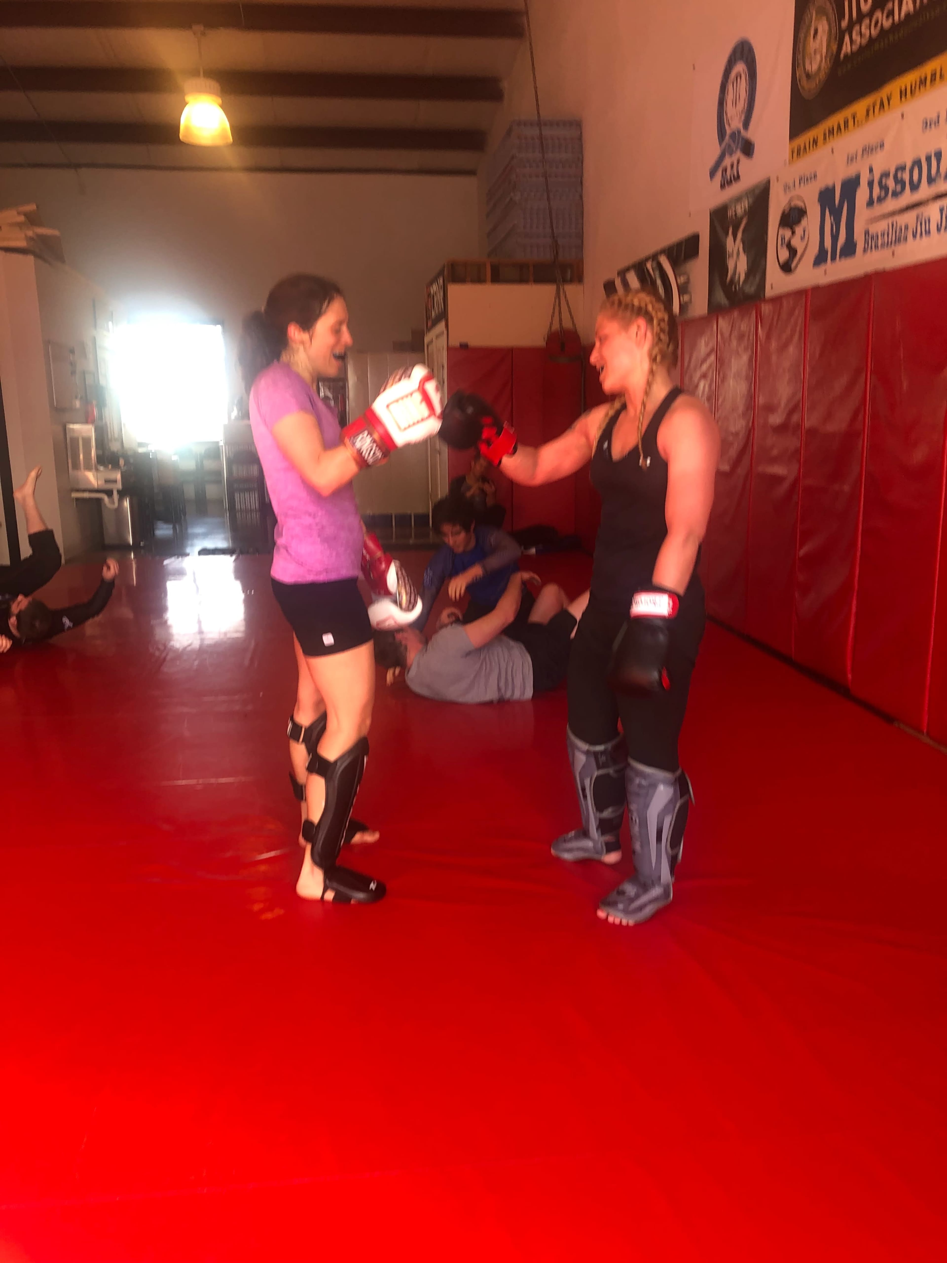 Two women in a martial arts gym wear sparring gear and practice boxing techniques while others sit on the mat in the background.