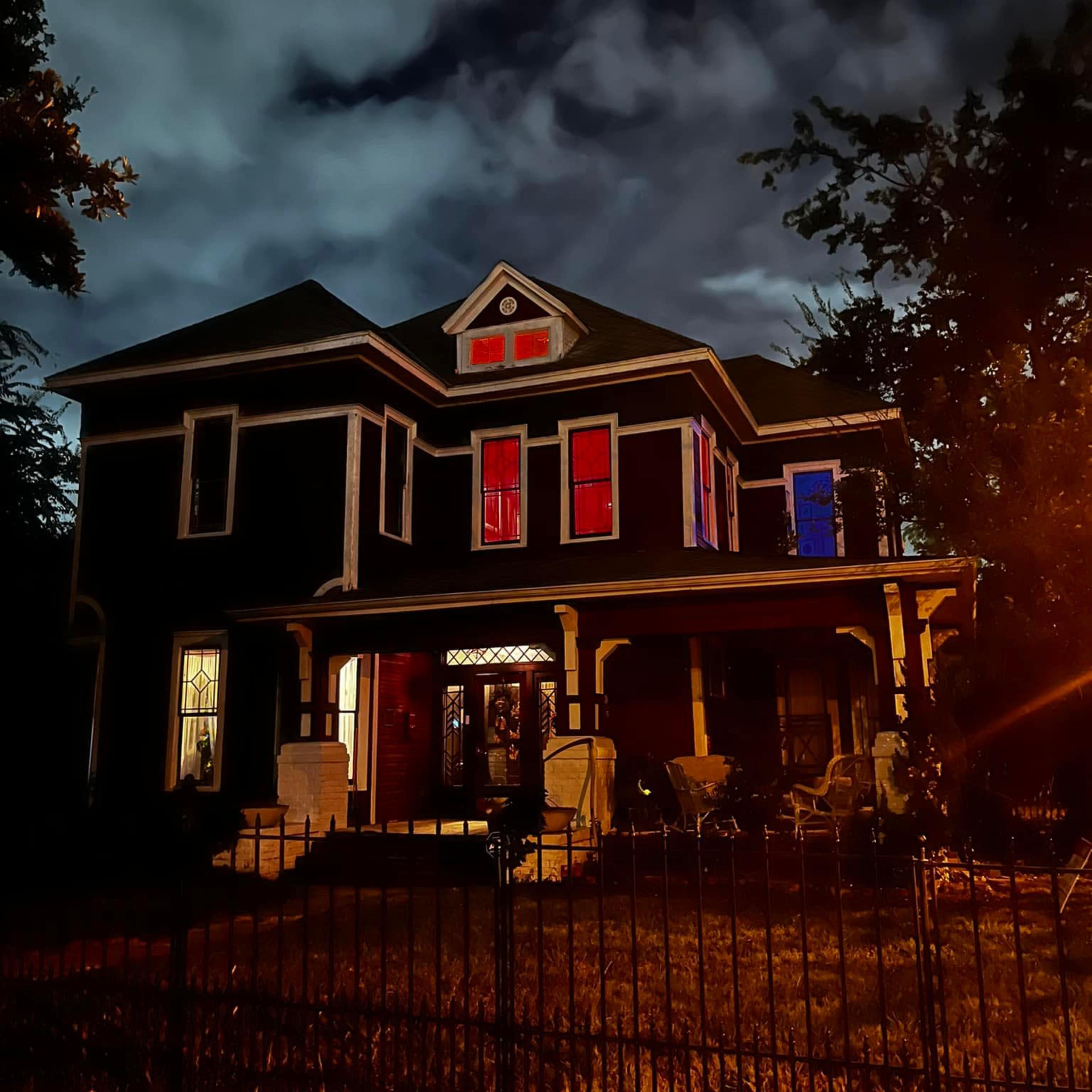 A dark, Victorian-style house with colorful lights in the windows and a wrought-iron fence is illuminated against a cloudy night sky.