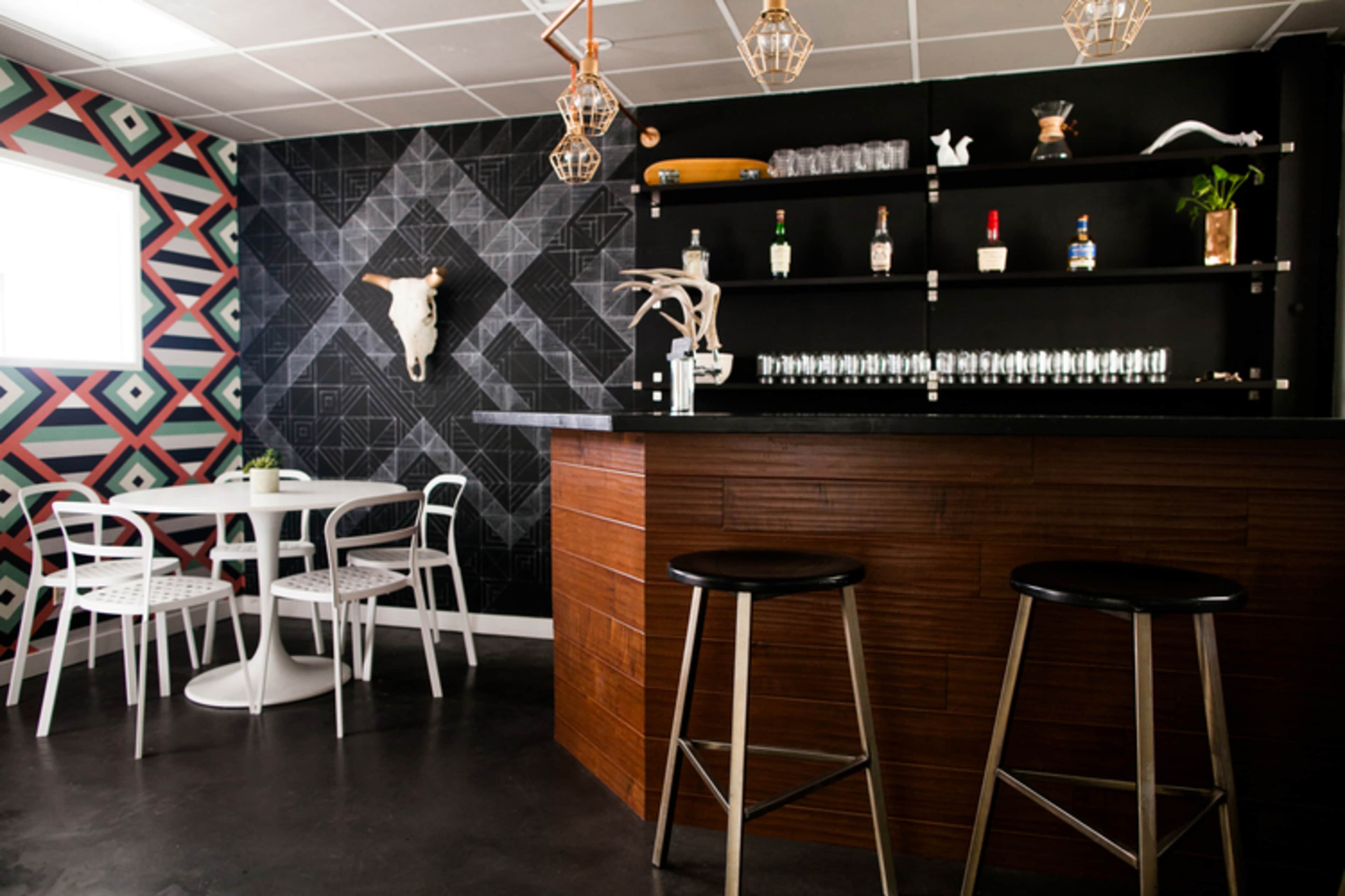 A modern bar area with a dark wooden counter, a minimalist round table with white chairs, and a geometric patterned wall adorned with a decorative skull.