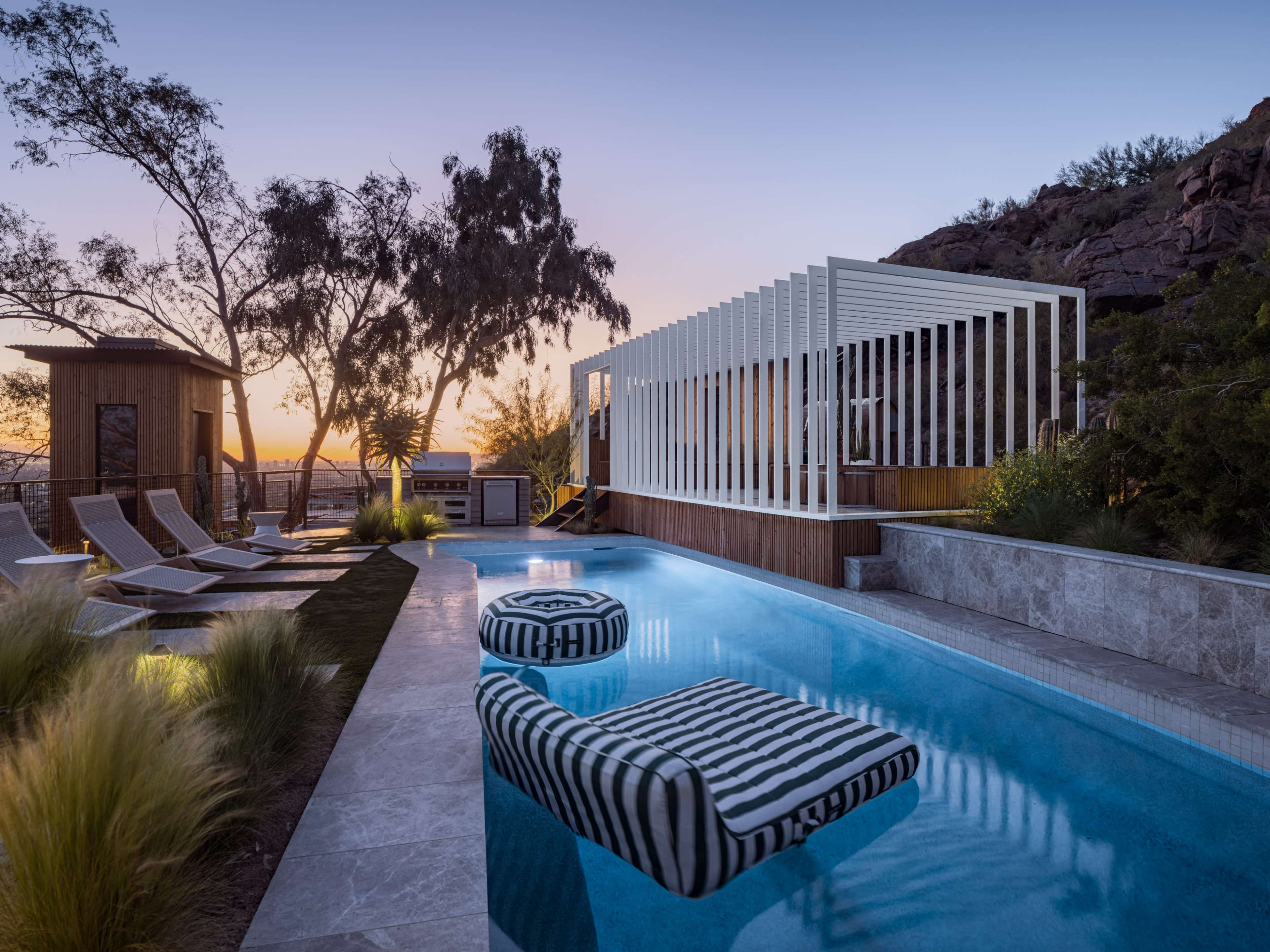 A modern outdoor pool area with striped lounge chairs, a white slatted structure, and a sunset view over a rocky landscape.