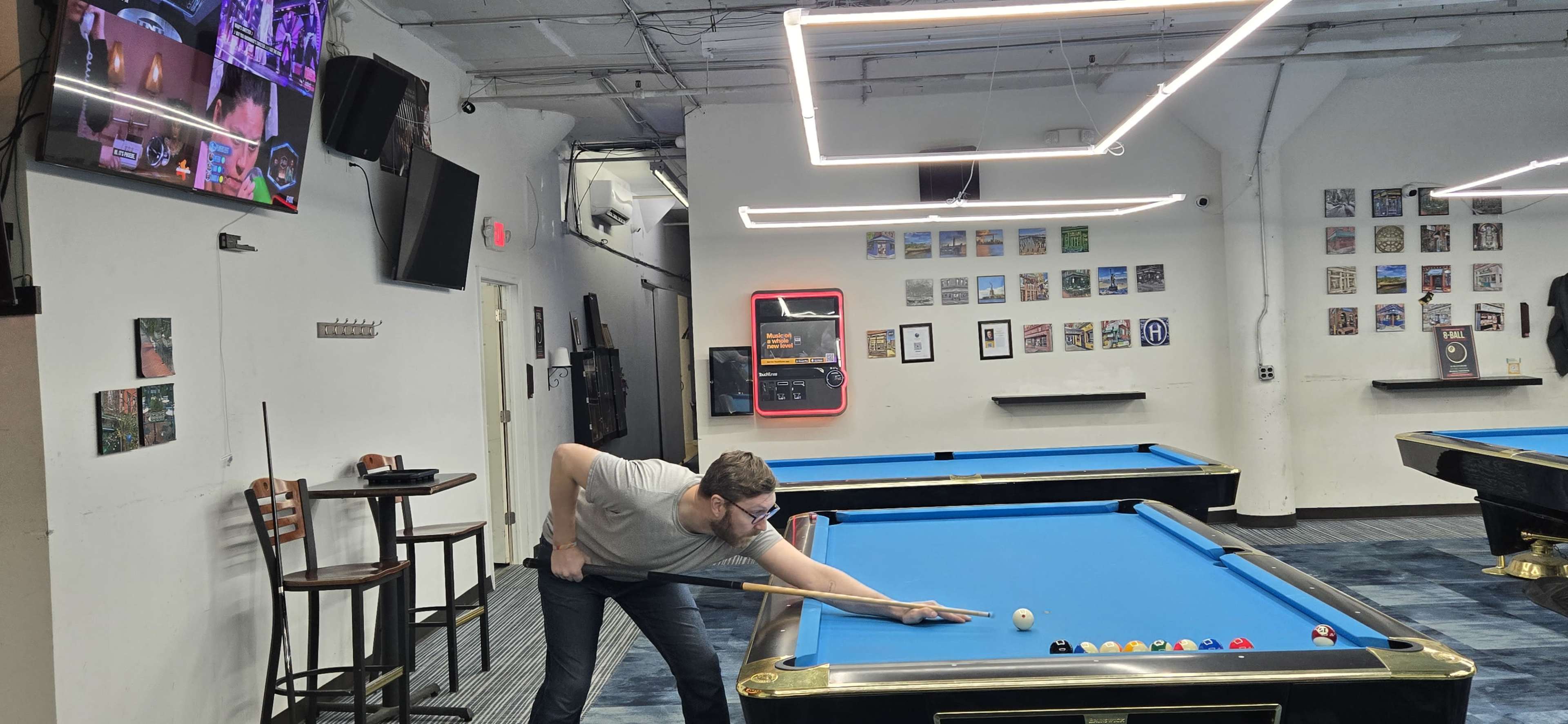 A man leans over a blue pool table aiming to strike the cue ball, with various gaming machines and artwork visible in the background.