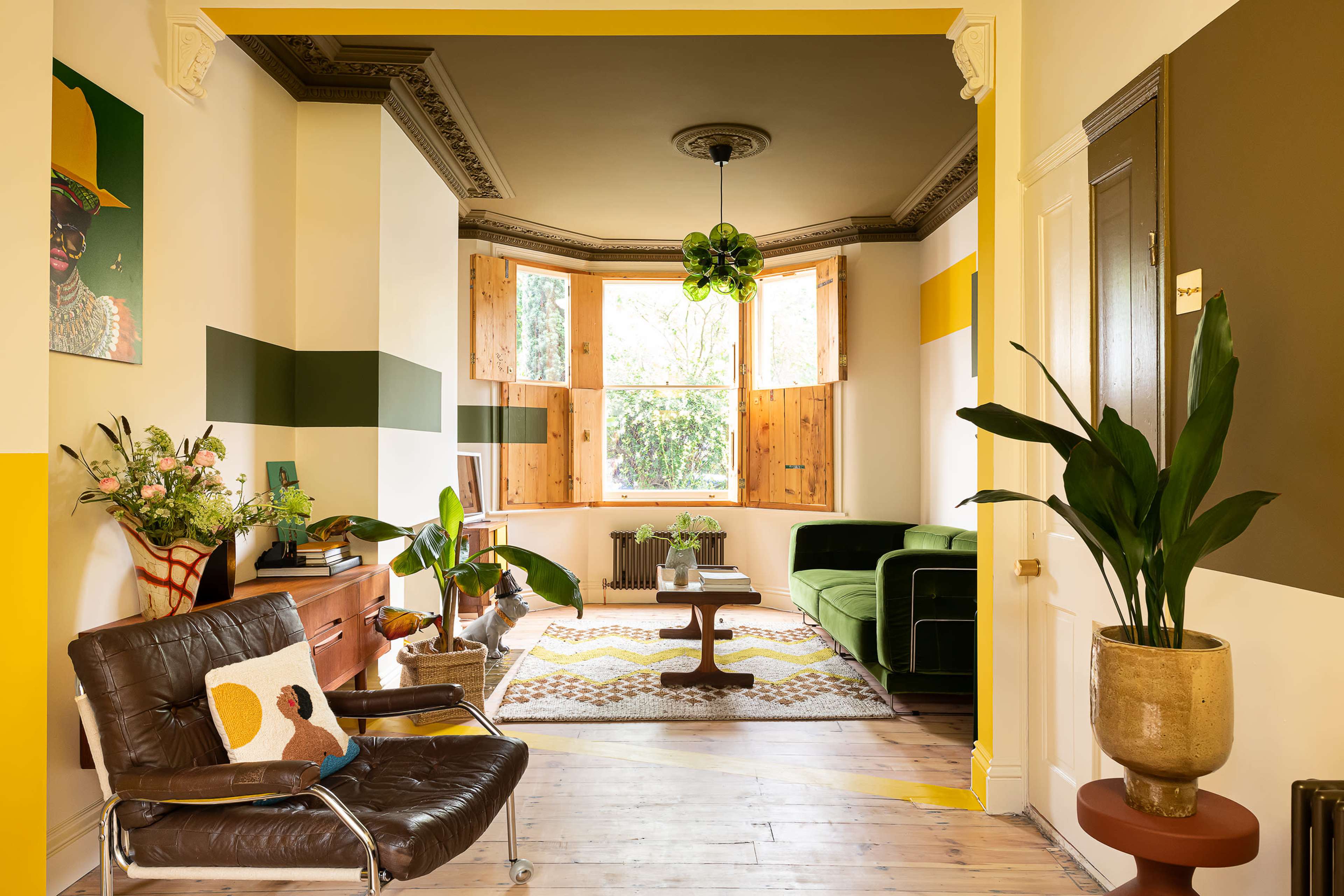 A brightly lit living room featuring a green sofa, a patterned rug, and large windows with wooden shutters.
