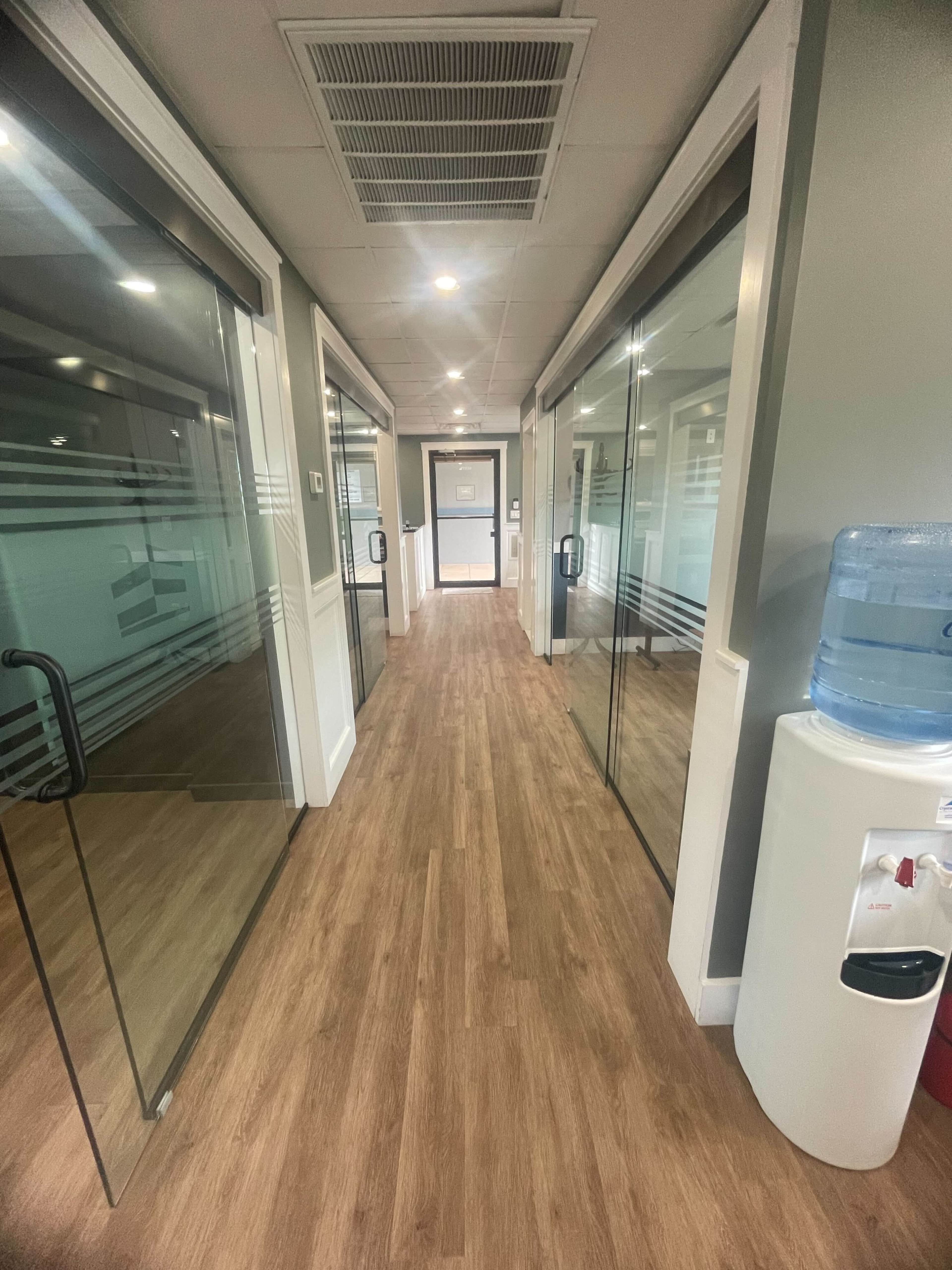 The image shows a narrow hallway with glass-walled offices on either side and a water cooler at the end.