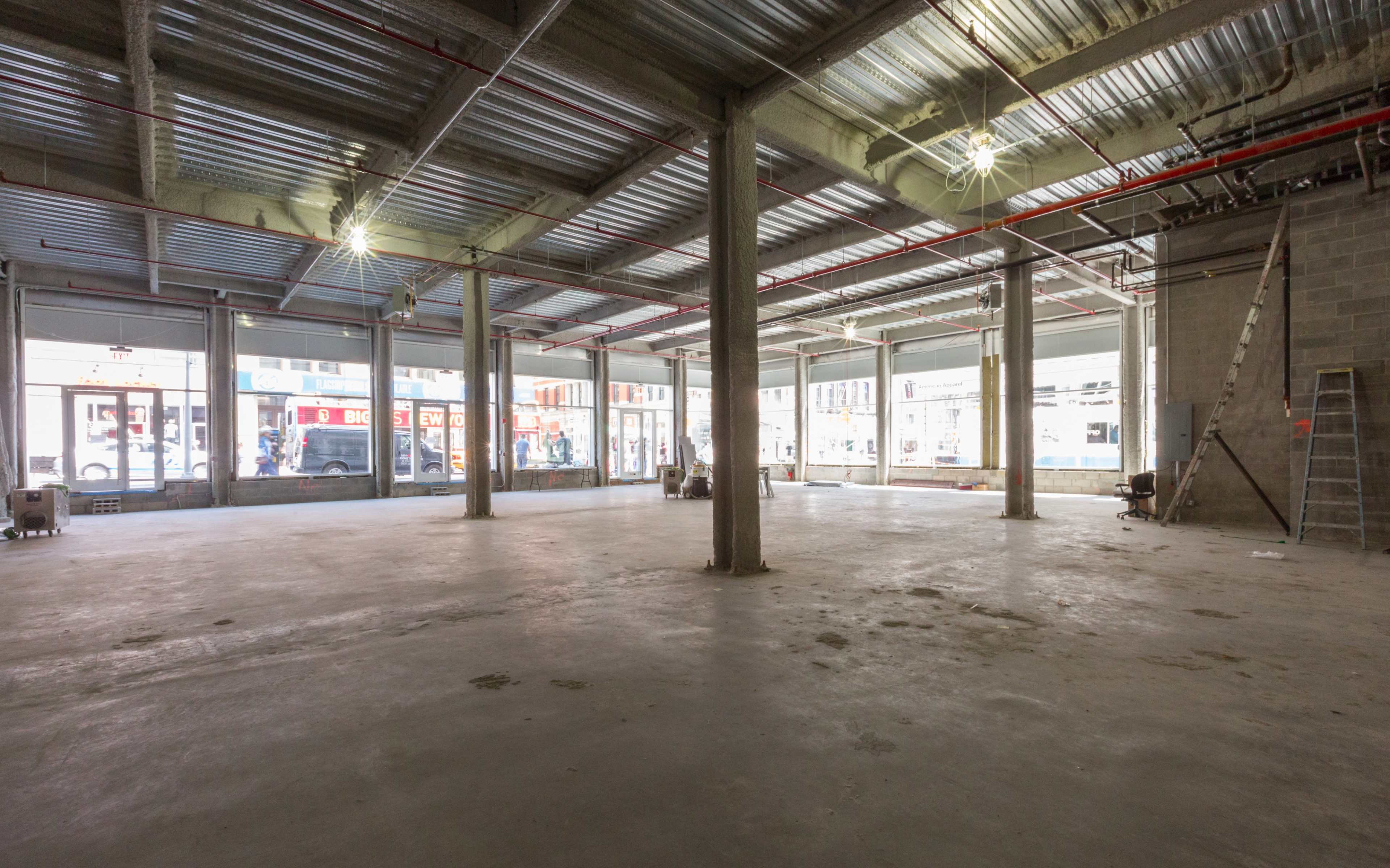 An empty commercial space with concrete flooring, exposed beams, and large windows letting in light.