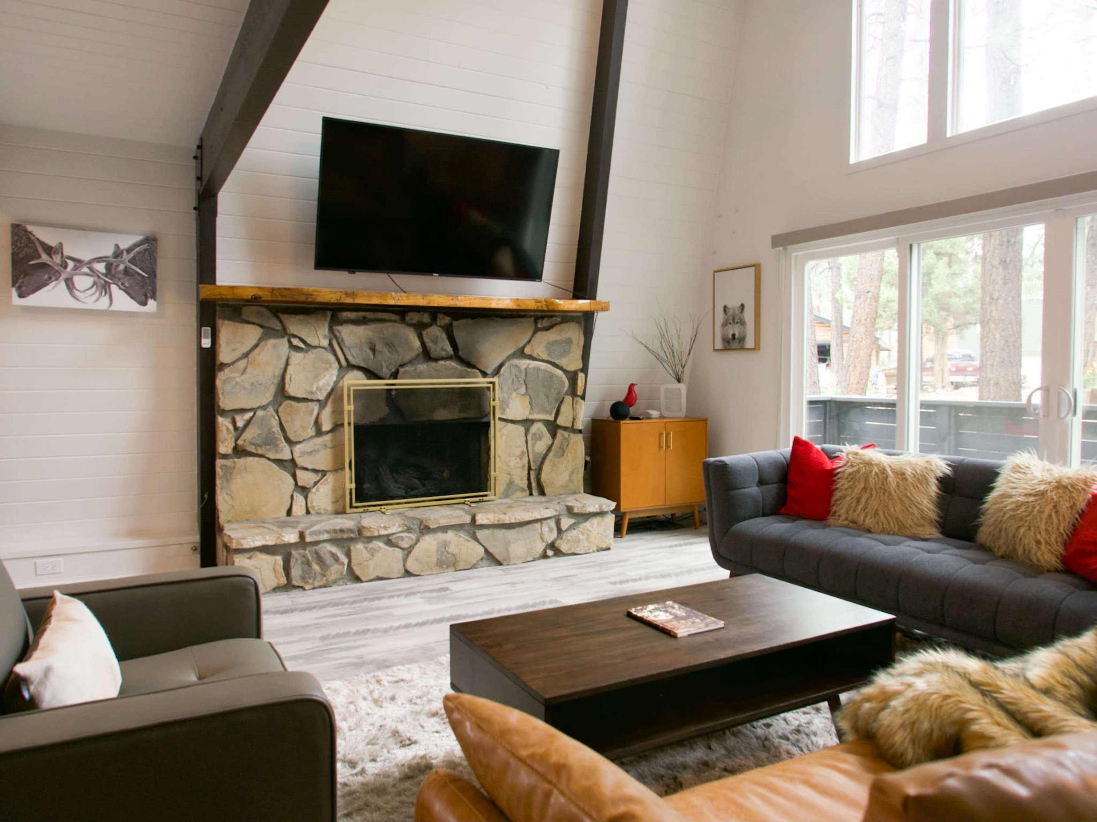 A modern living room with a stone fireplace, a large television mounted above it, and a coffee table surrounded by dark furniture and decorative pillows.