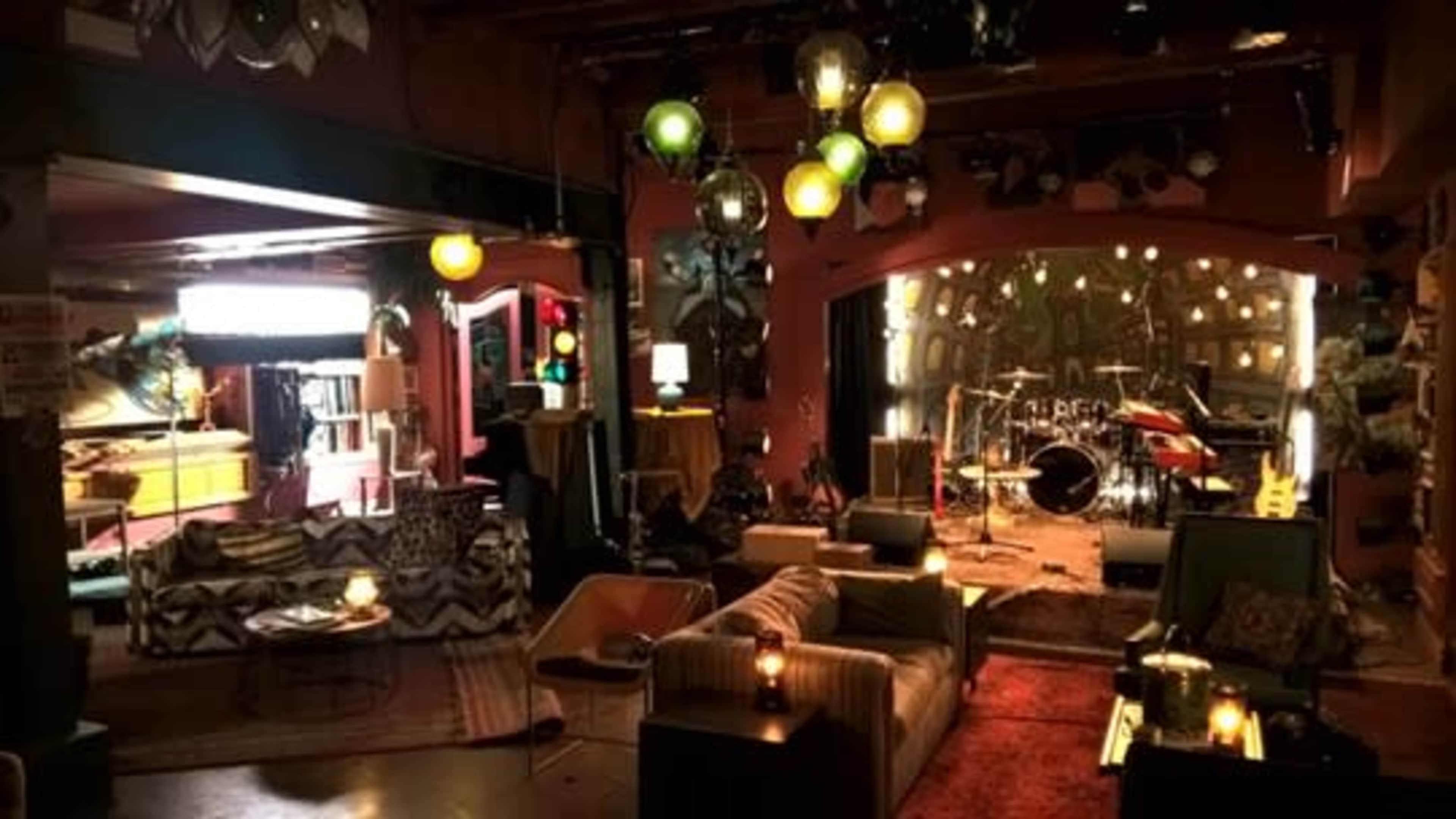 The image shows a dimly lit music venue with a stage set up for a live performance, surrounded by cozy seating areas and decorative lighting.