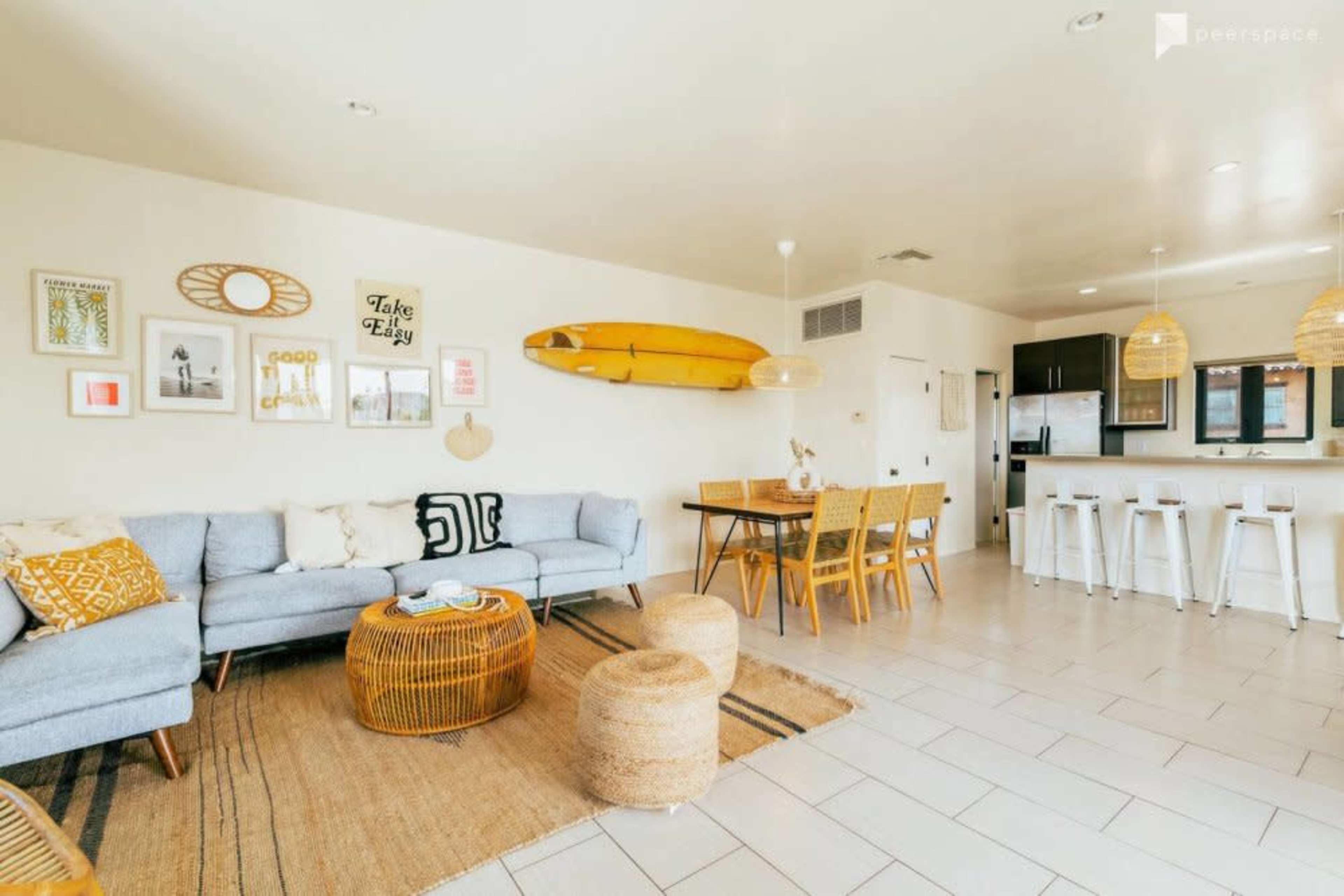 The image shows a modern living space featuring a light-colored sofa, a round rattan coffee table, dining chairs, a dining table, and a kitchen area with a surfboard mounted on the wall.