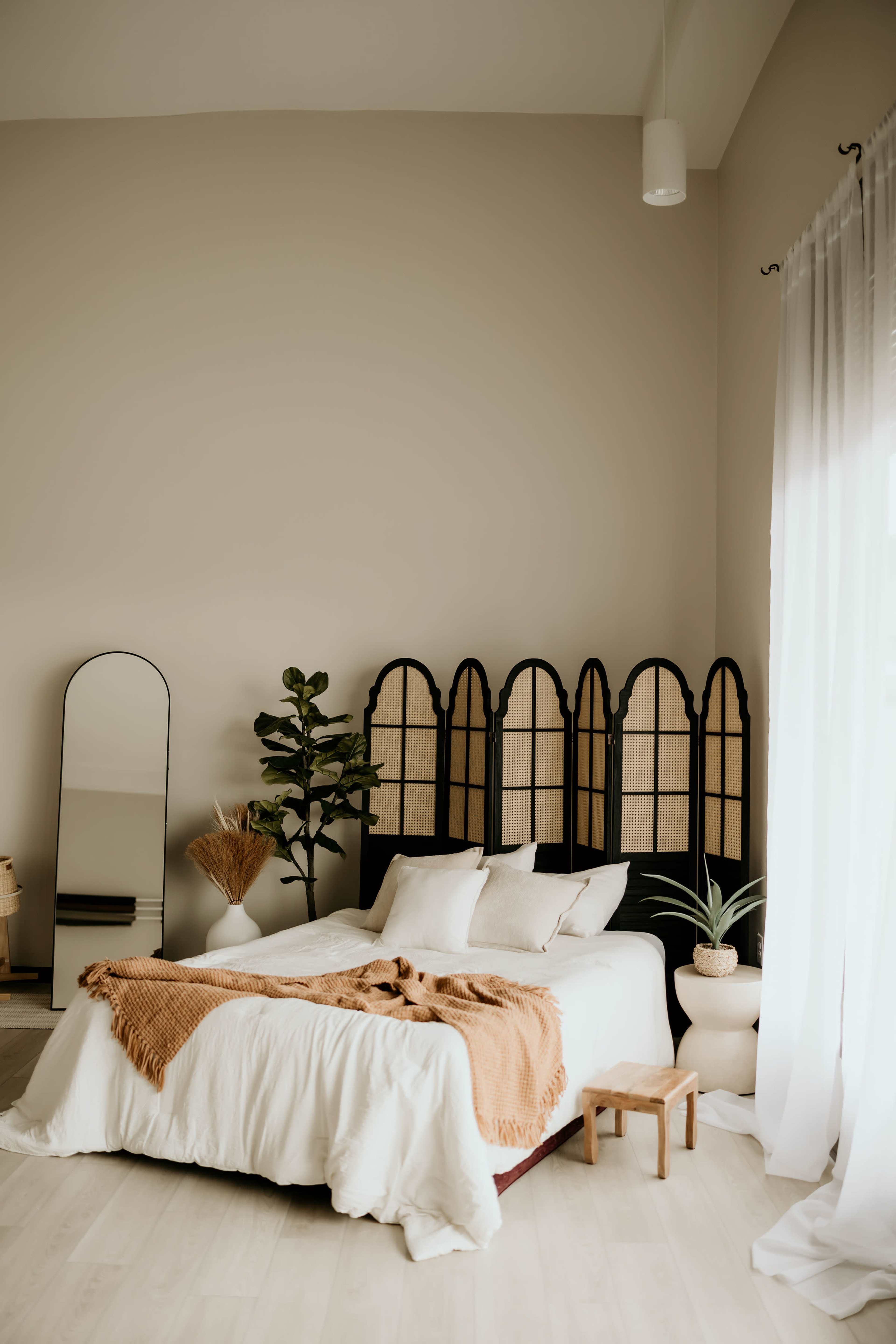 A bright bedroom features a bed with white bedding, a folding screen behind it, a mirror, and potted plants by the window.