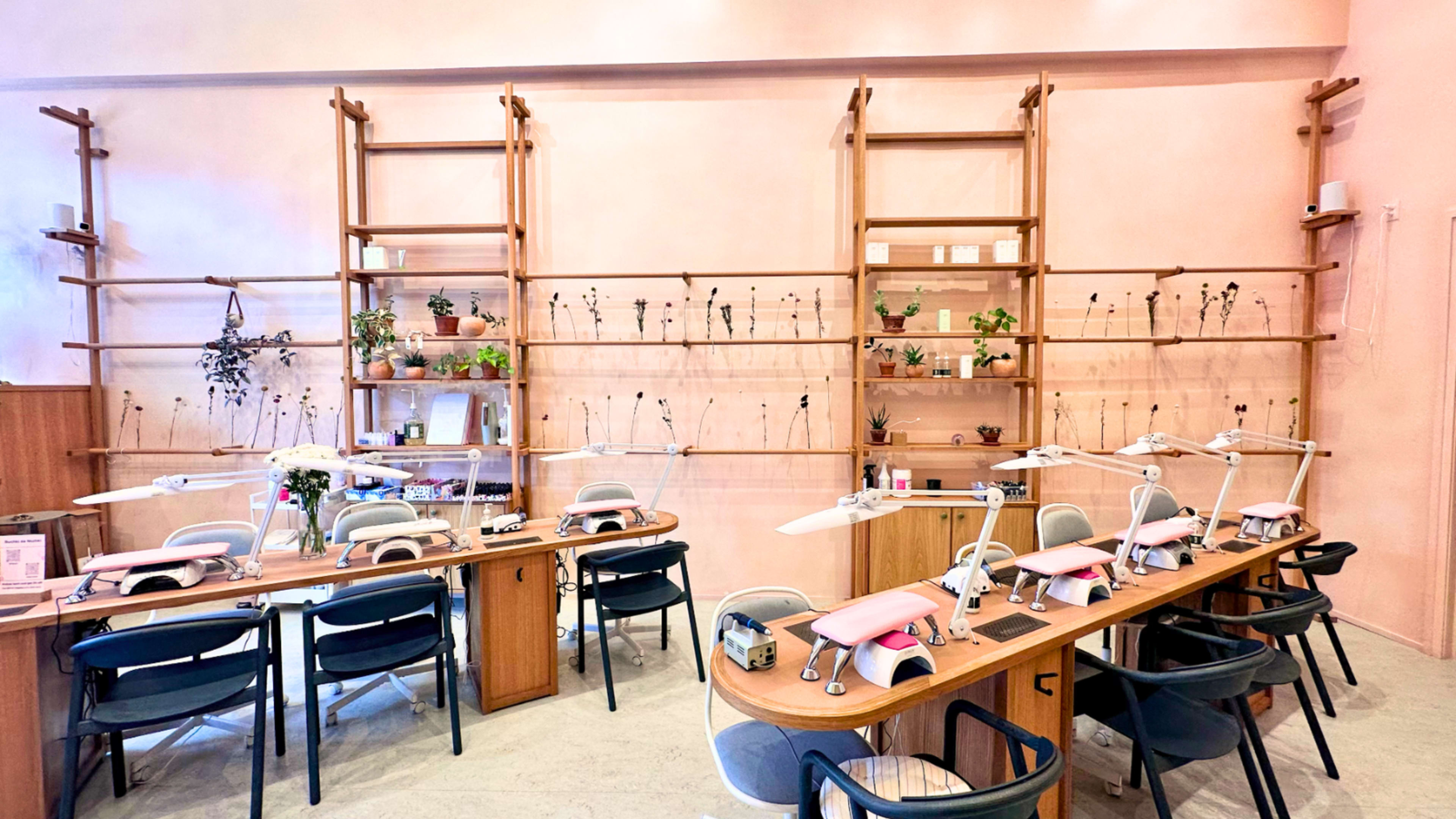 The image shows a modern nail salon with wooden tables and chairs arranged neatly, featuring plants on shelves and workstations equipped with nail care tools.