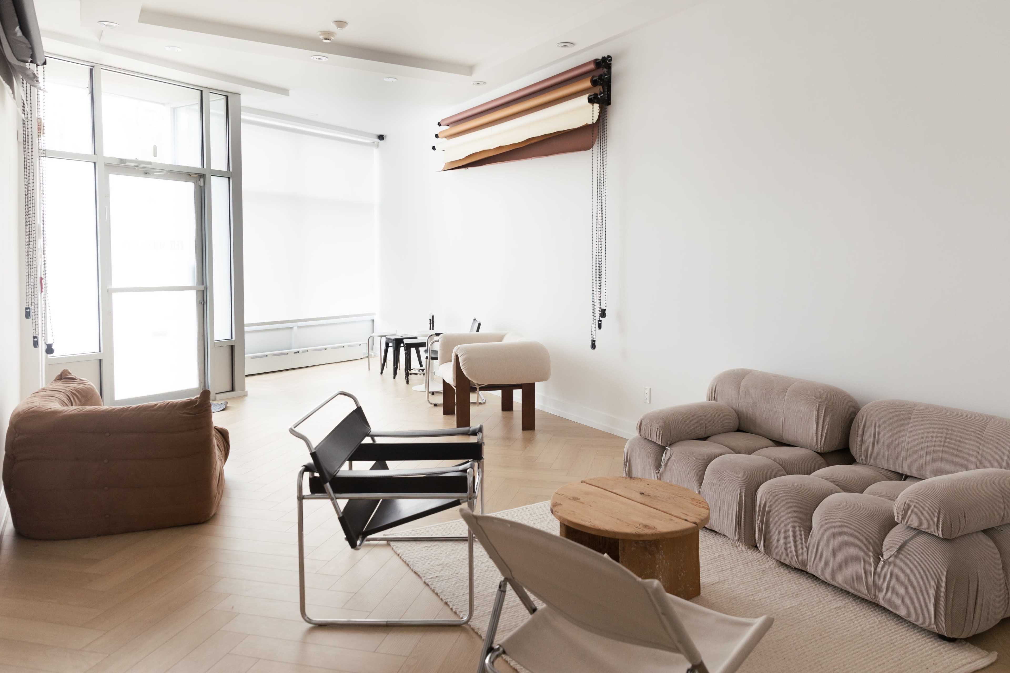The spacious, minimalist room features various seating options, including a sectional sofa, lounge chairs, and a wooden table, all arranged around light-colored wooden flooring.