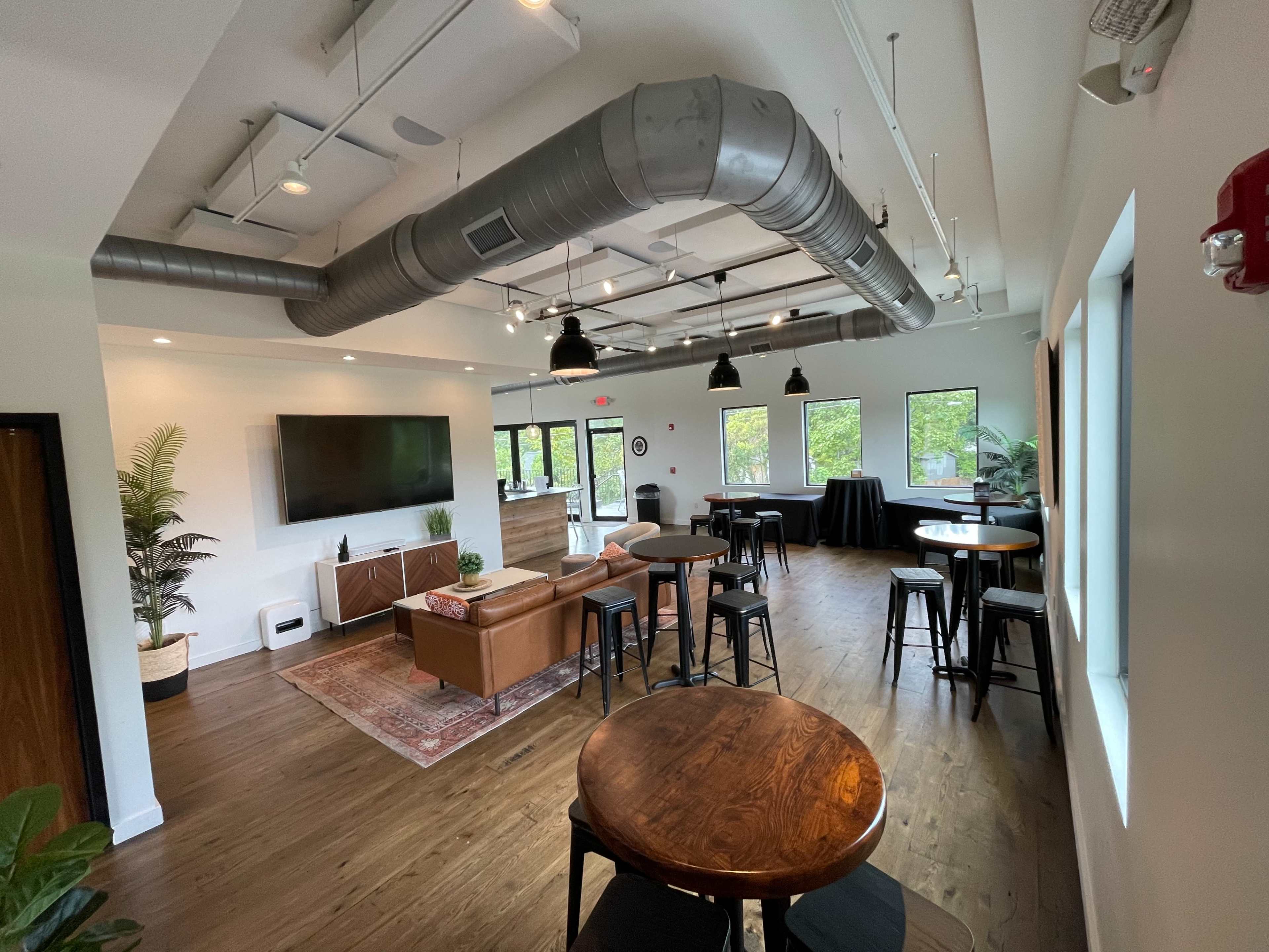 A modern lounge area with wooden floors, several tables, black chairs, a large television on the wall, and a sofa.
