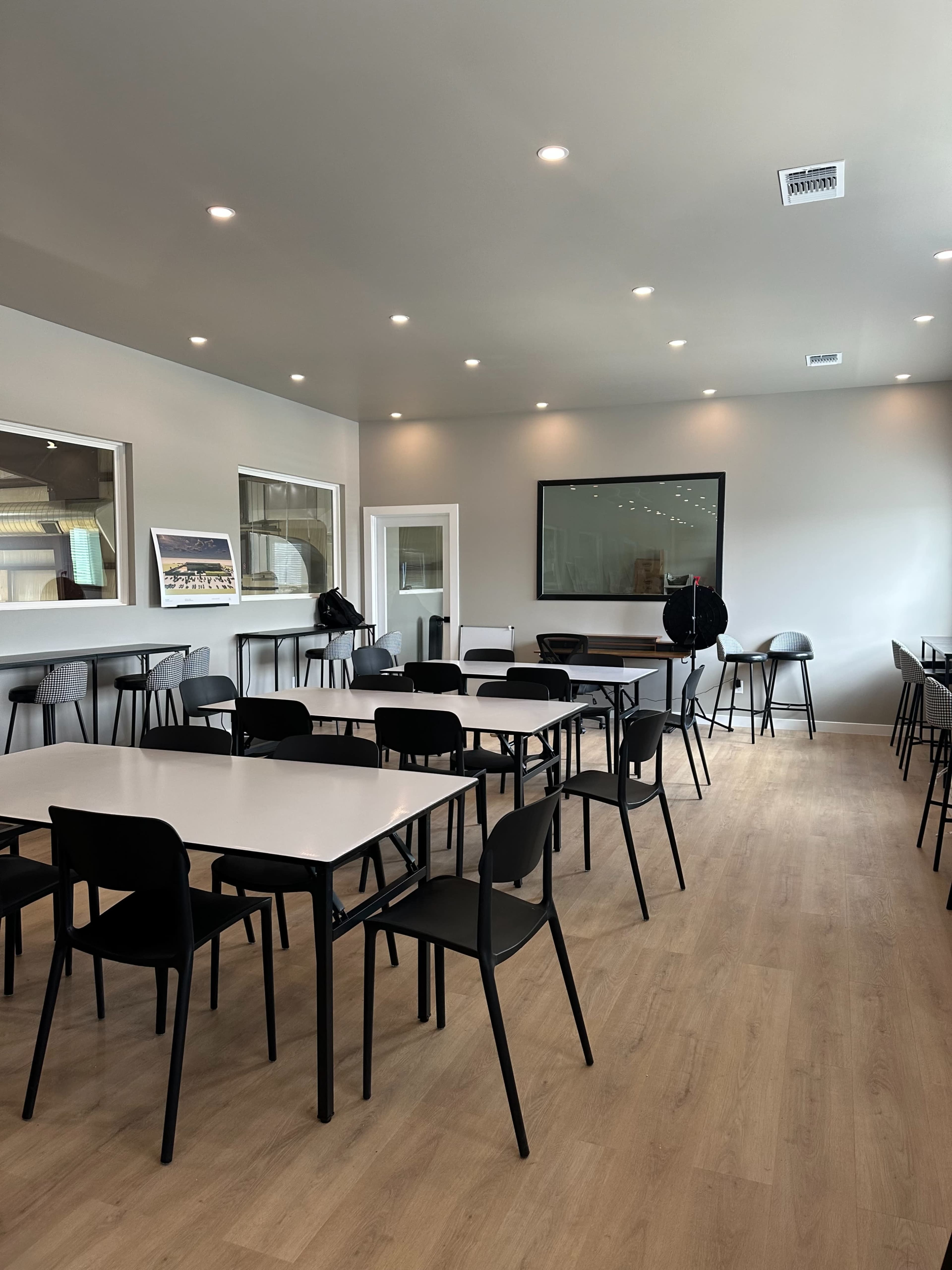 The image shows a modern, spacious meeting room with multiple tables and chairs arranged around the space, brightened by ample overhead lighting.