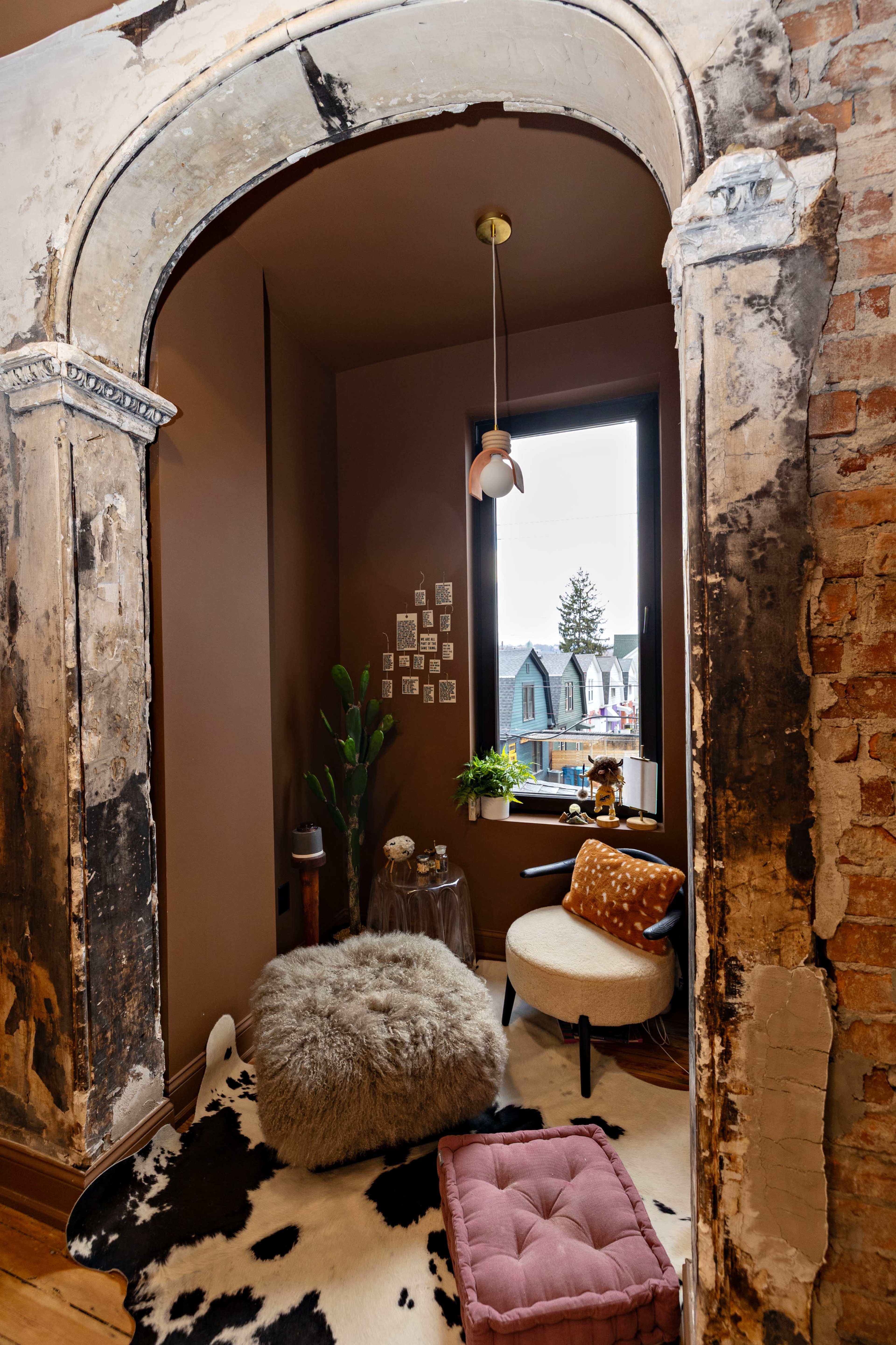 A cozy reading nook features a plush fur ottoman, a patterned chair, and a large window framed by an archway with exposed brick and plaster.