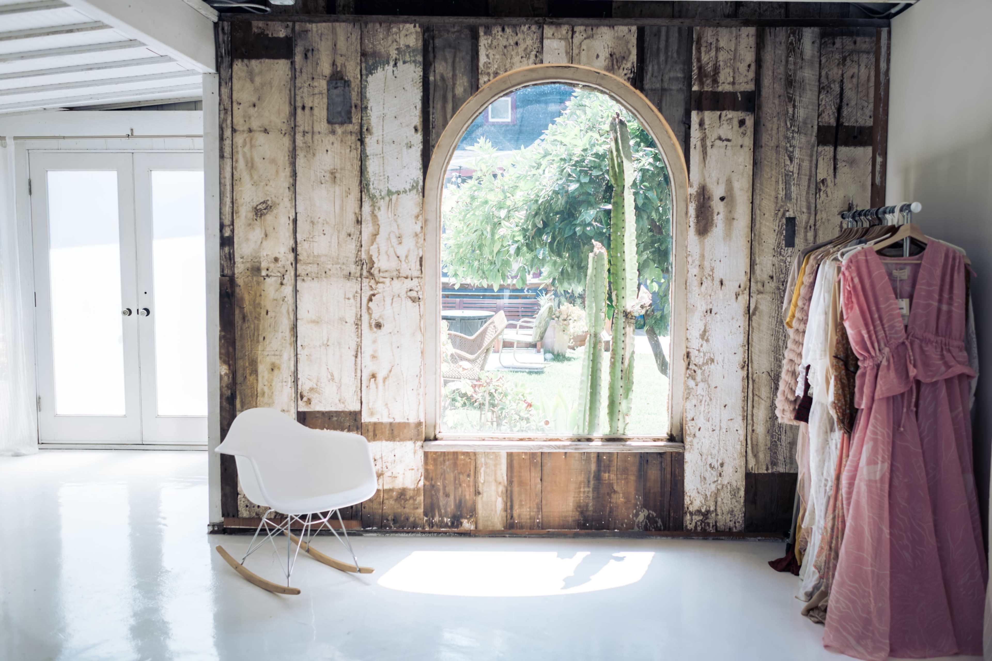 A sunlit room features a wooden arched window overlooking a garden, with a rocking chair and racks of clothing nearby.