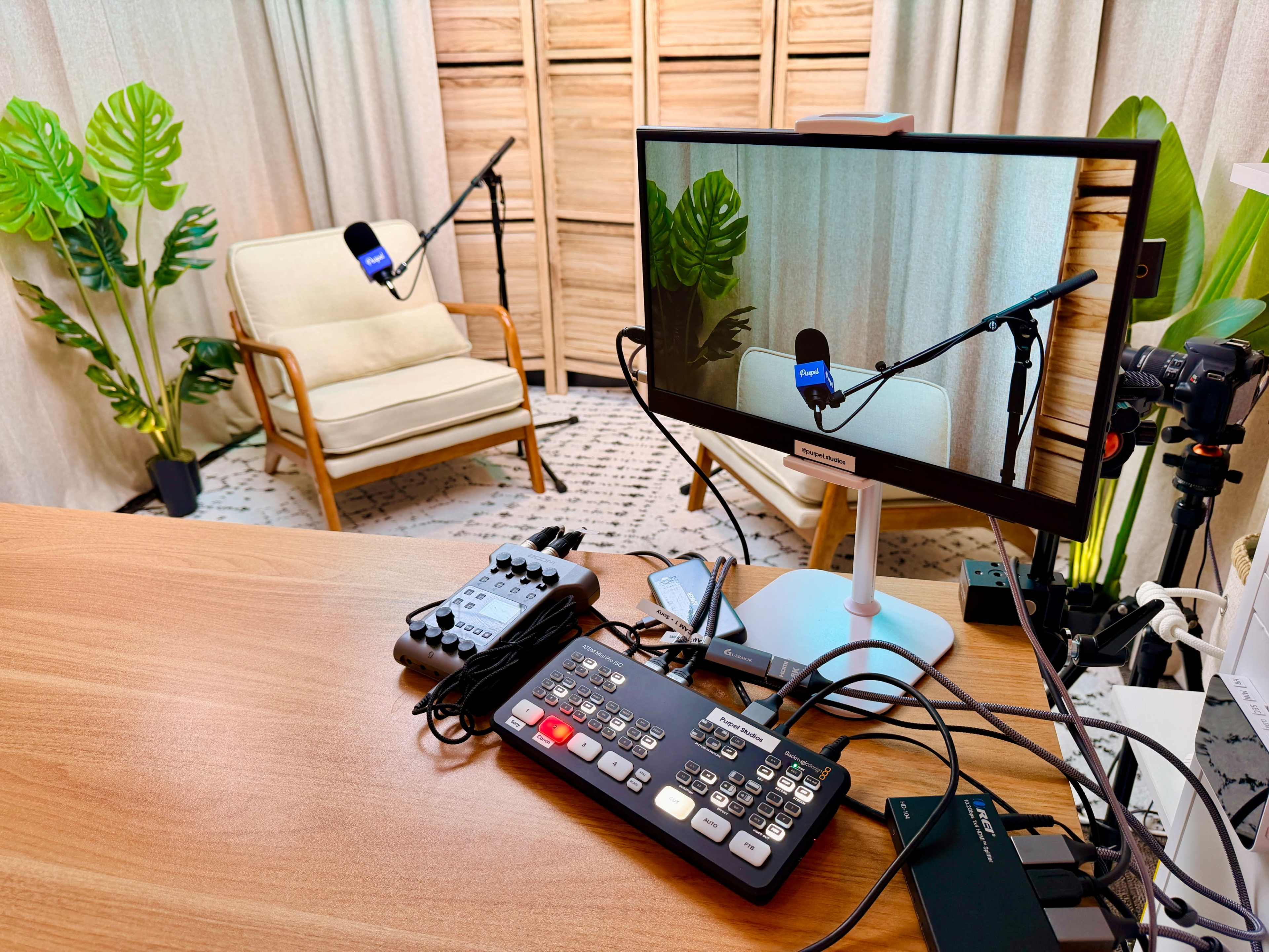 The image shows a recording setup featuring a chair, microphones, a computer monitor, and various equipment on a wooden desk, with potted plants in the background.