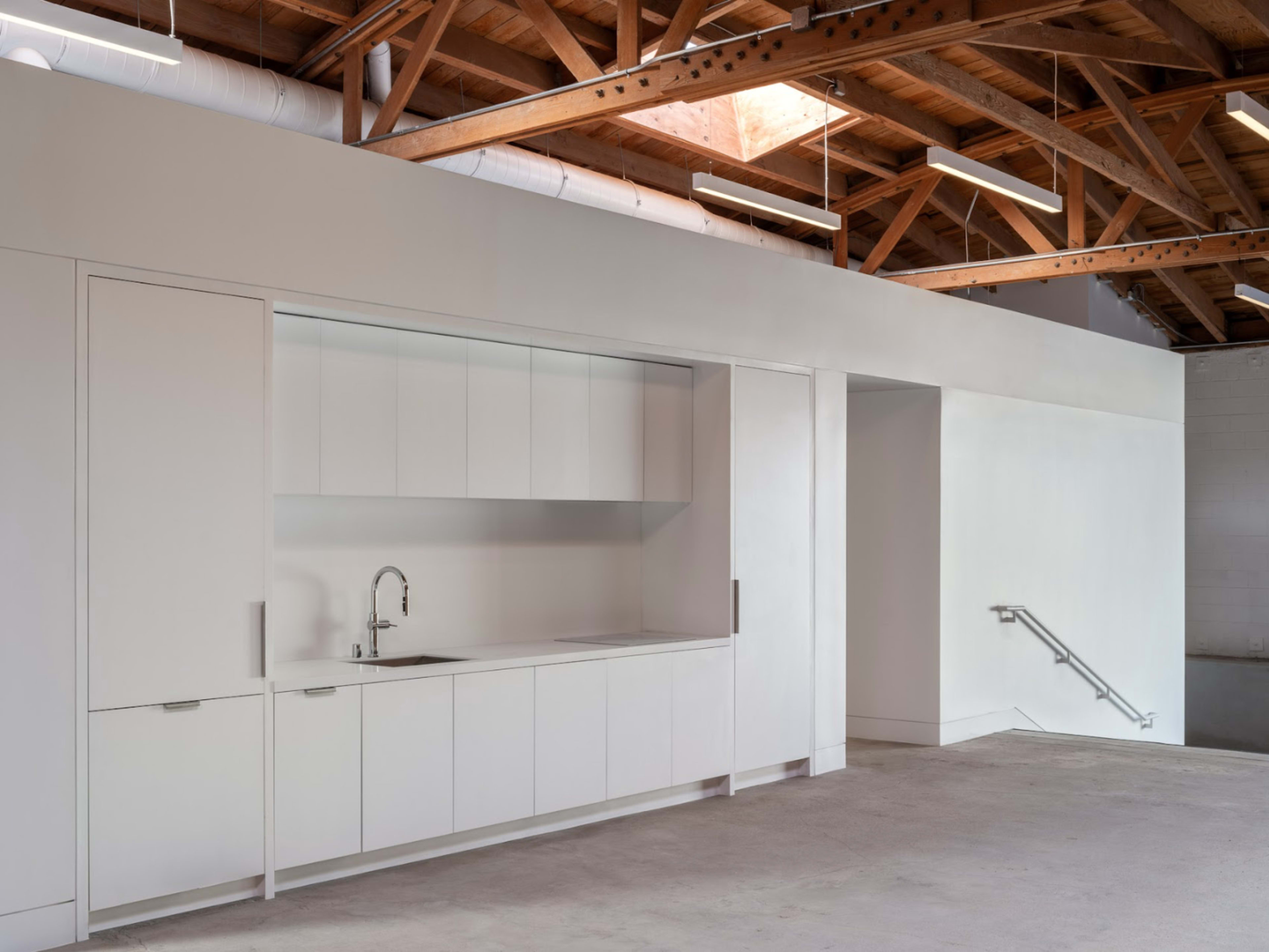 A modern kitchen space with a minimalistic white cabinetry setup against a pale wall and exposed wooden beams above.