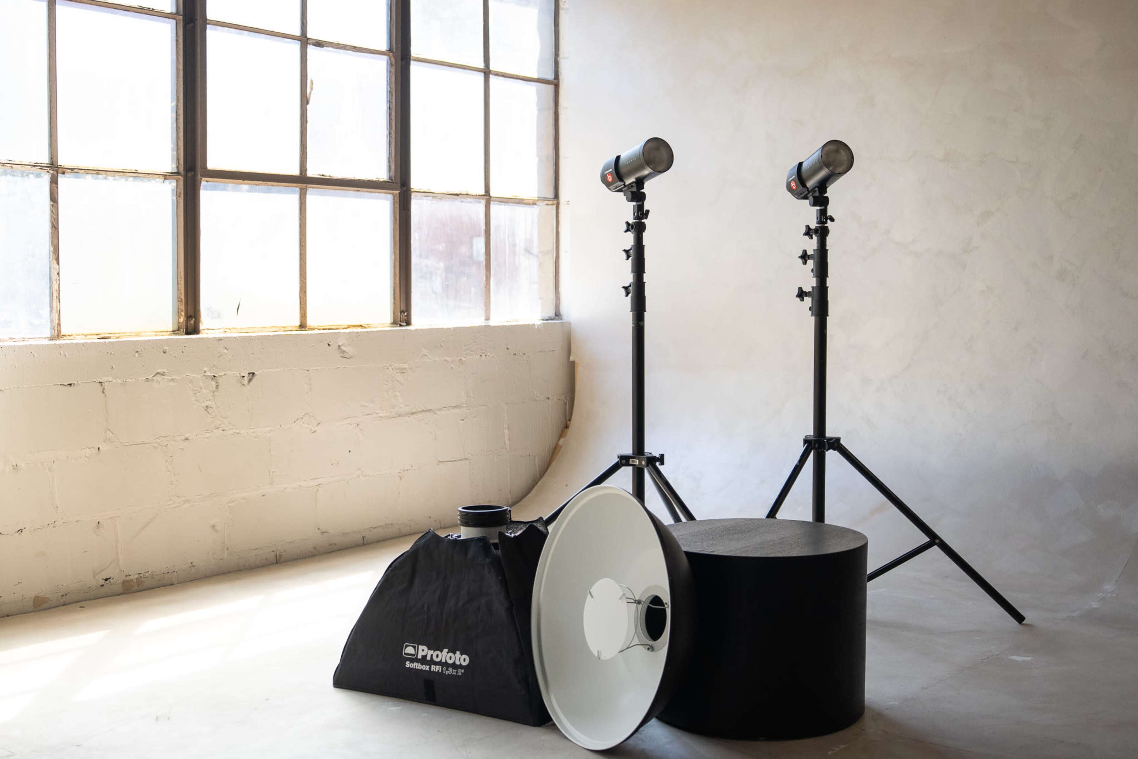 The image shows two studio light fixtures on stands positioned near a large window in a minimalist indoor space.