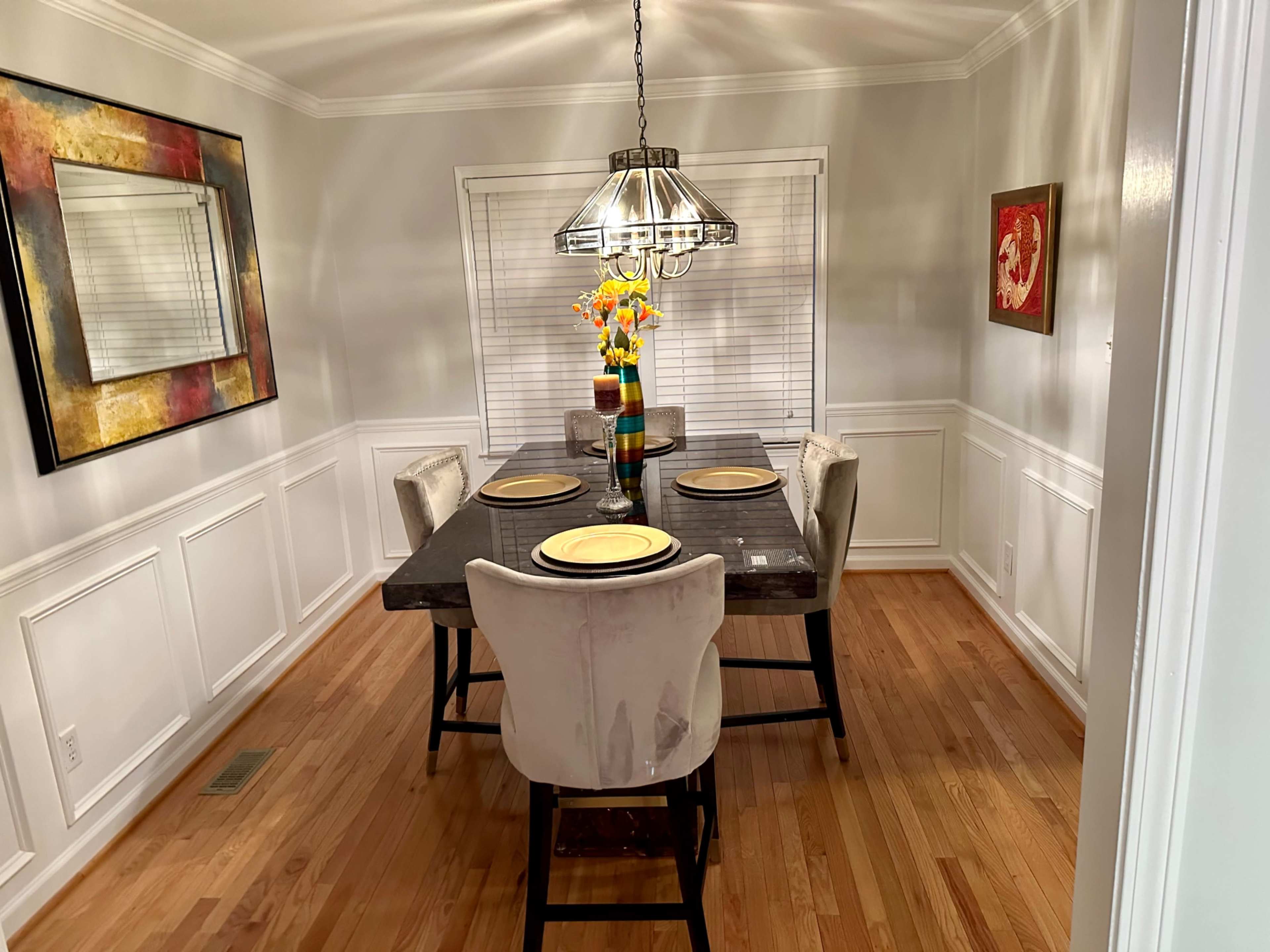 A dining room features a rectangular table set for four, adorned with a floral centerpiece, surrounded by upholstered chairs and illuminated by a hanging light fixture.