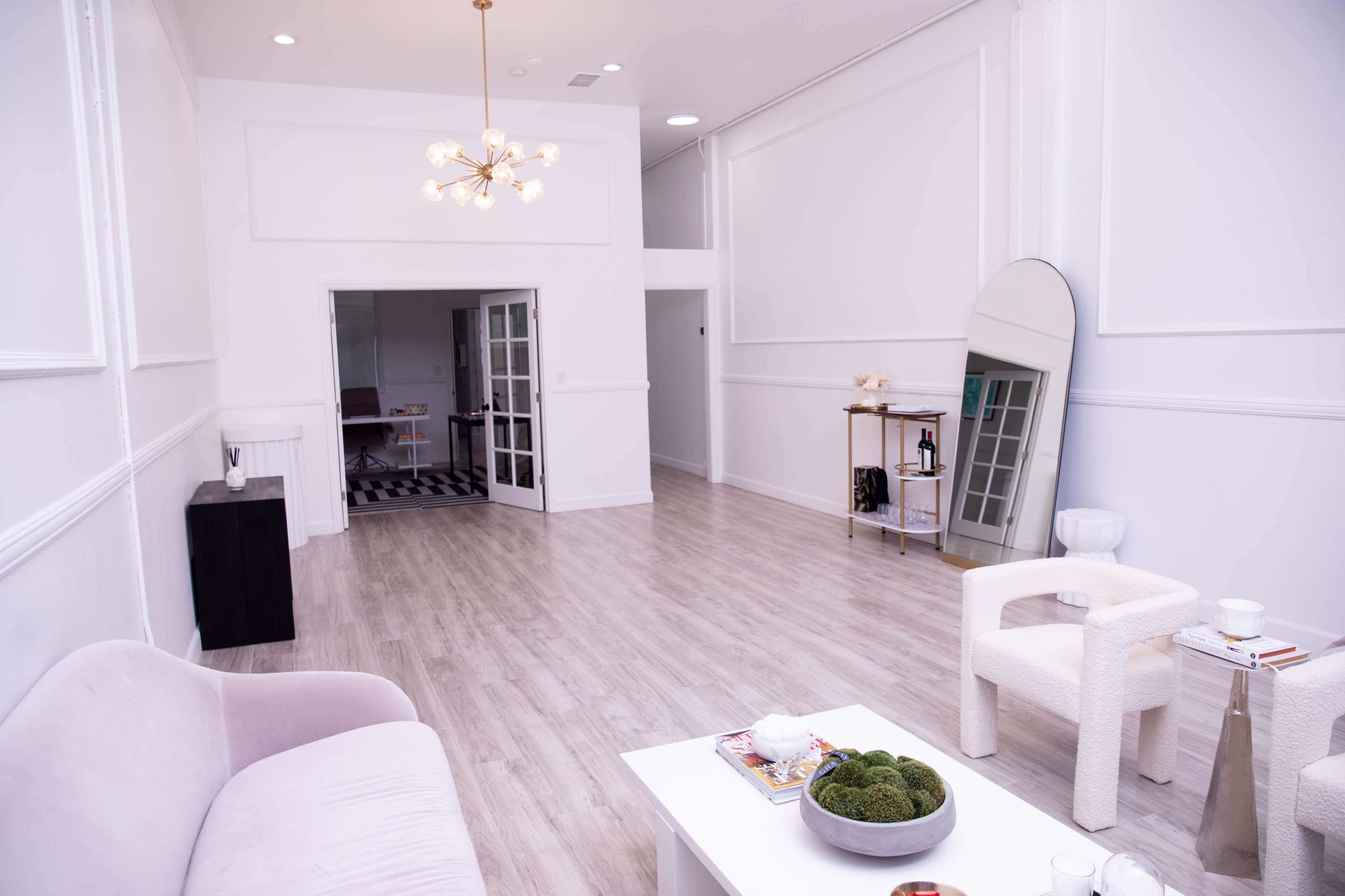 A spacious, minimalistic room with light wooden flooring, featuring a pink sofa, a white coffee table with decor, and an oversized mirror against the wall.