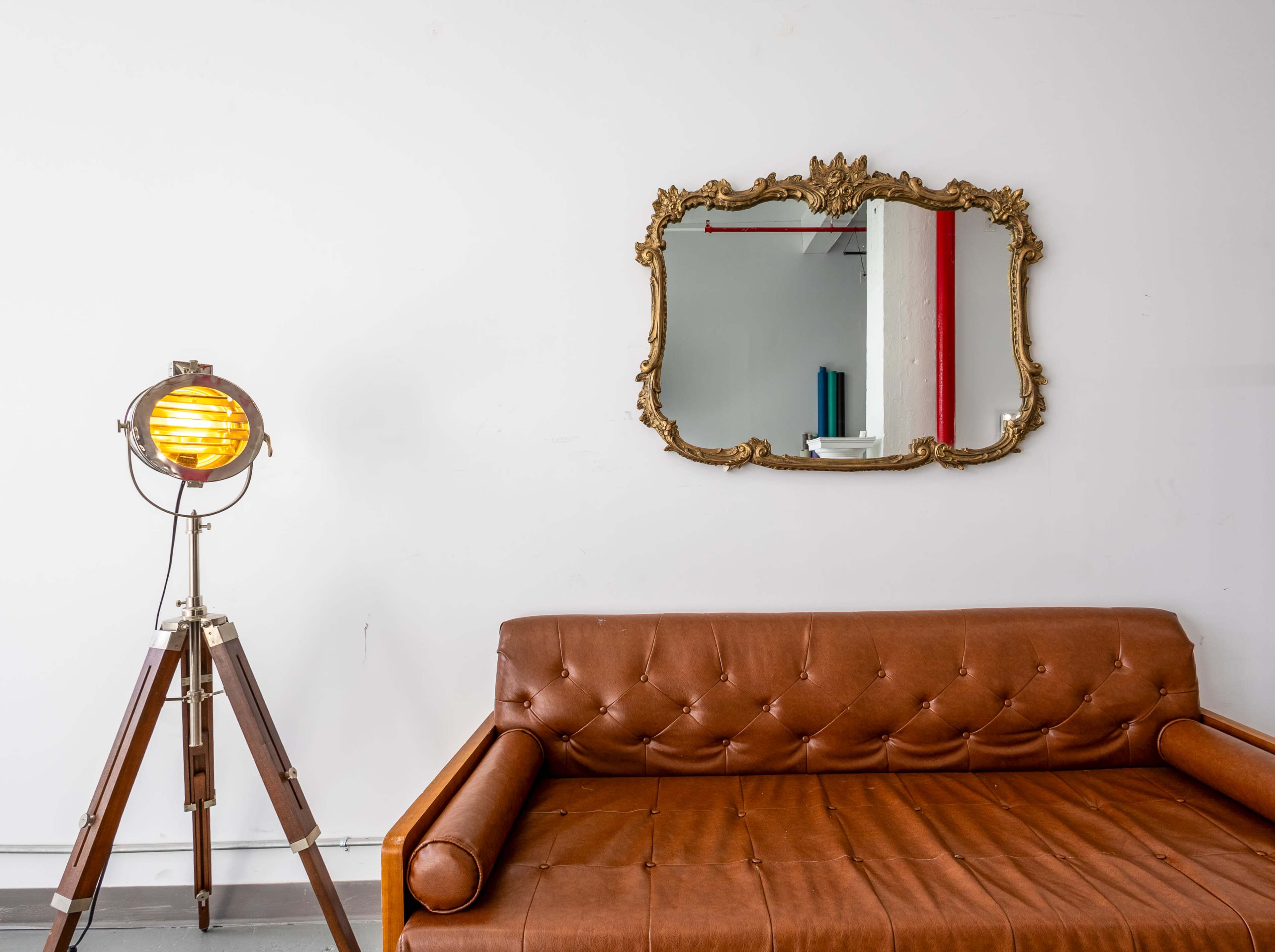 A brown leather sofa is positioned against a white wall, accompanied by a vintage-style lamp and an ornate mirror.