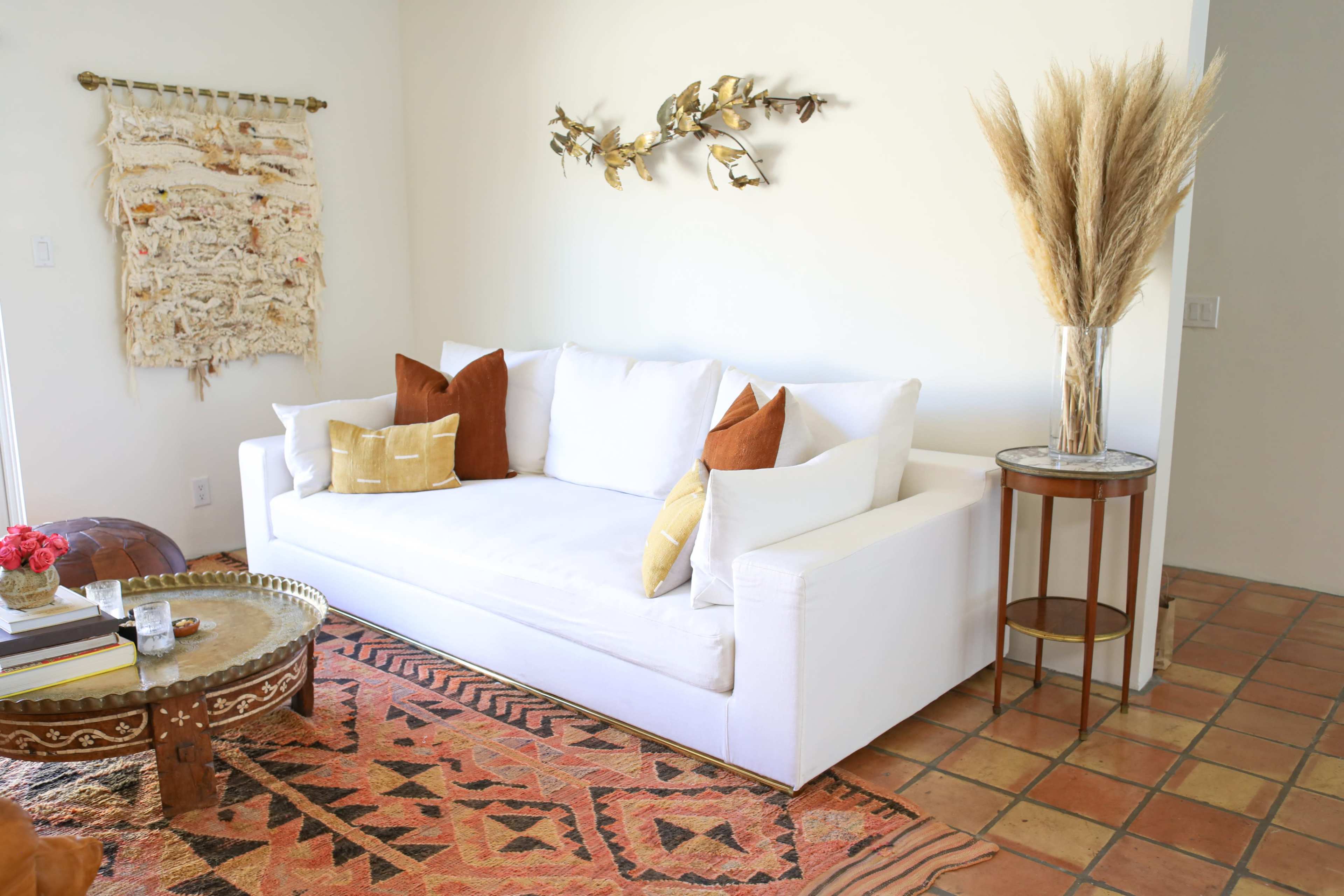 A modern living room features a white sofa with decorative pillows, a patterned rug, a wooden coffee table, and a vase of pampas grass on a side table.