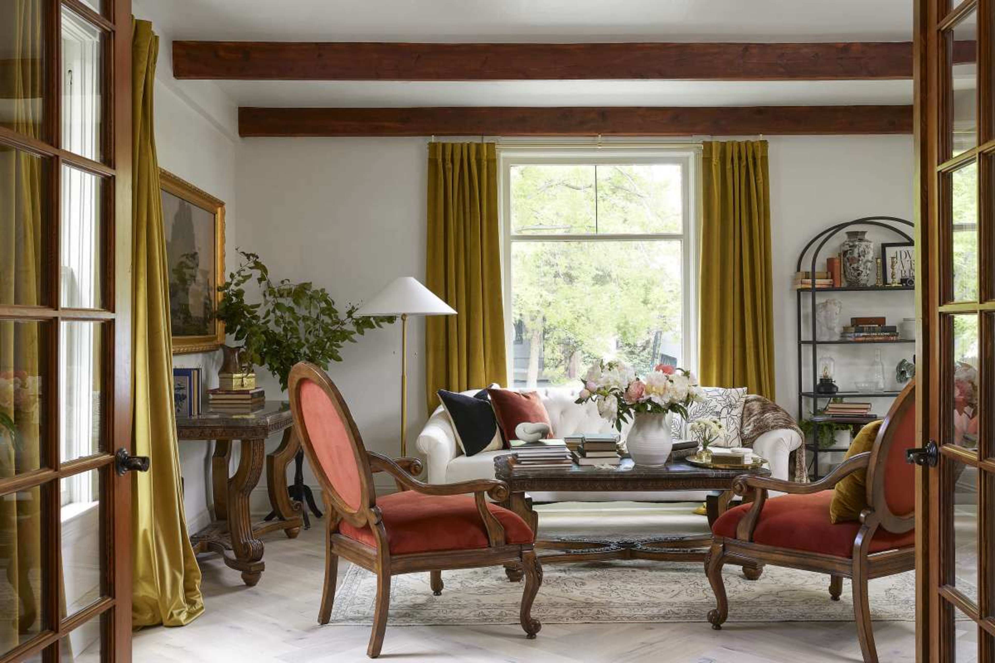A stylish living room features a white sofa, two red upholstered chairs, a wooden coffee table, and large windows dressed with yellow curtains.