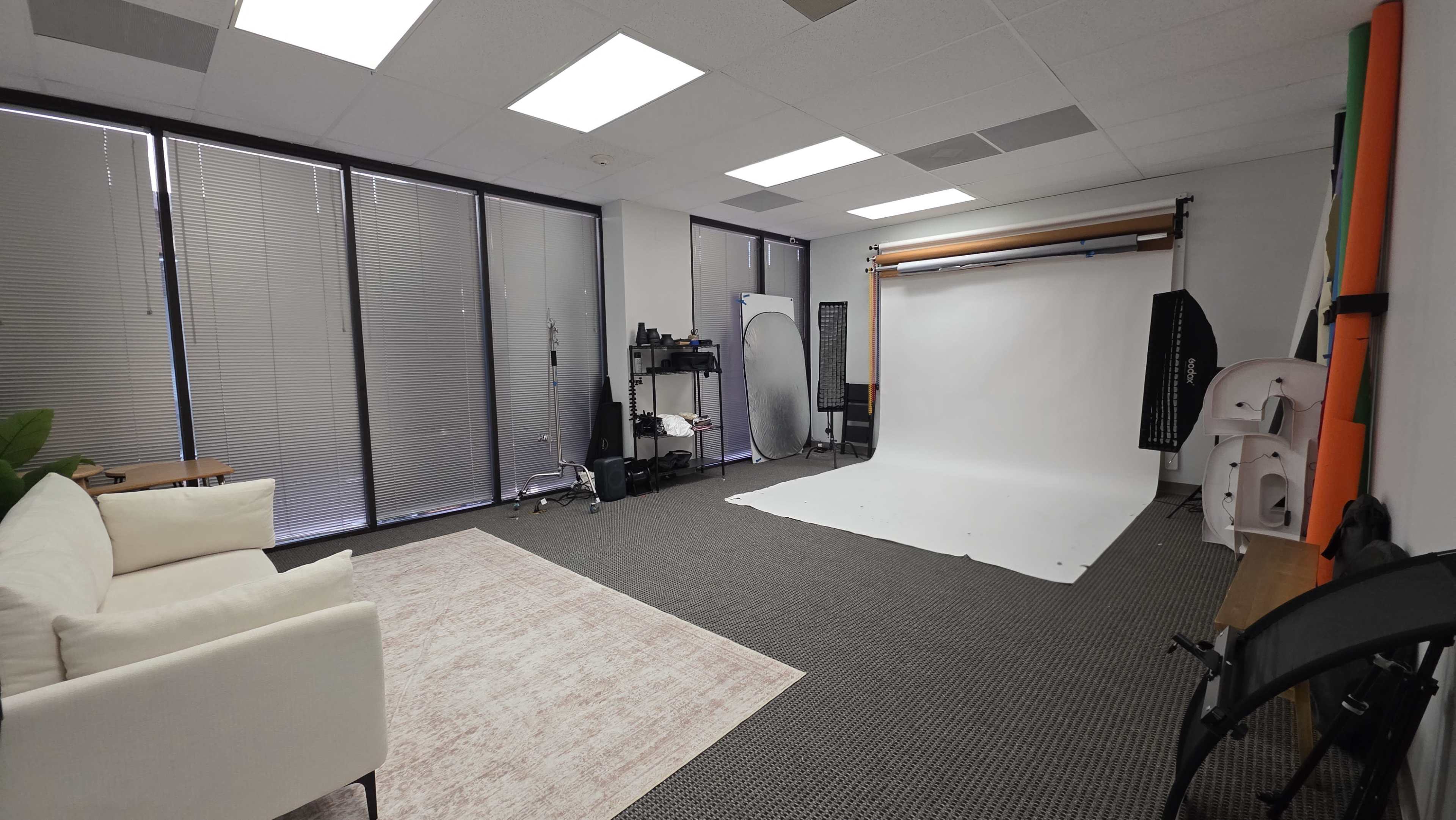 The image shows a spacious photography studio with a white backdrop, soft lighting equipment, and seating areas.