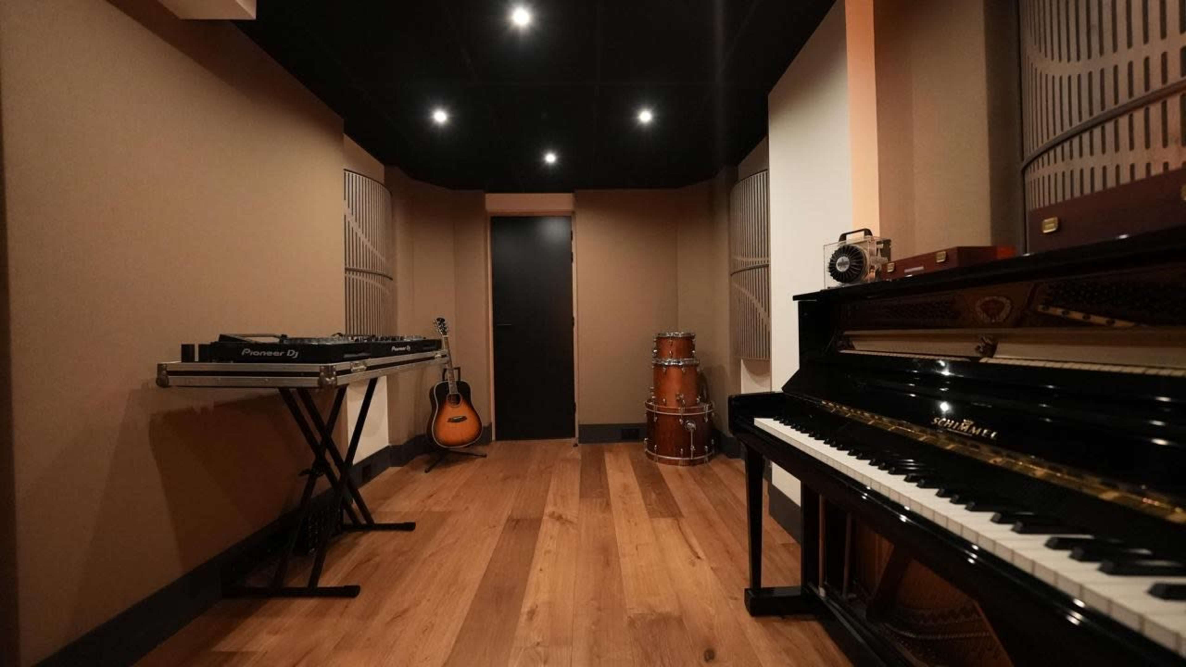 The image shows a music studio with a grand piano, an acoustic guitar, electronic equipment on a stand, and a conga drum in a well-lit, minimalist space with wooden flooring.