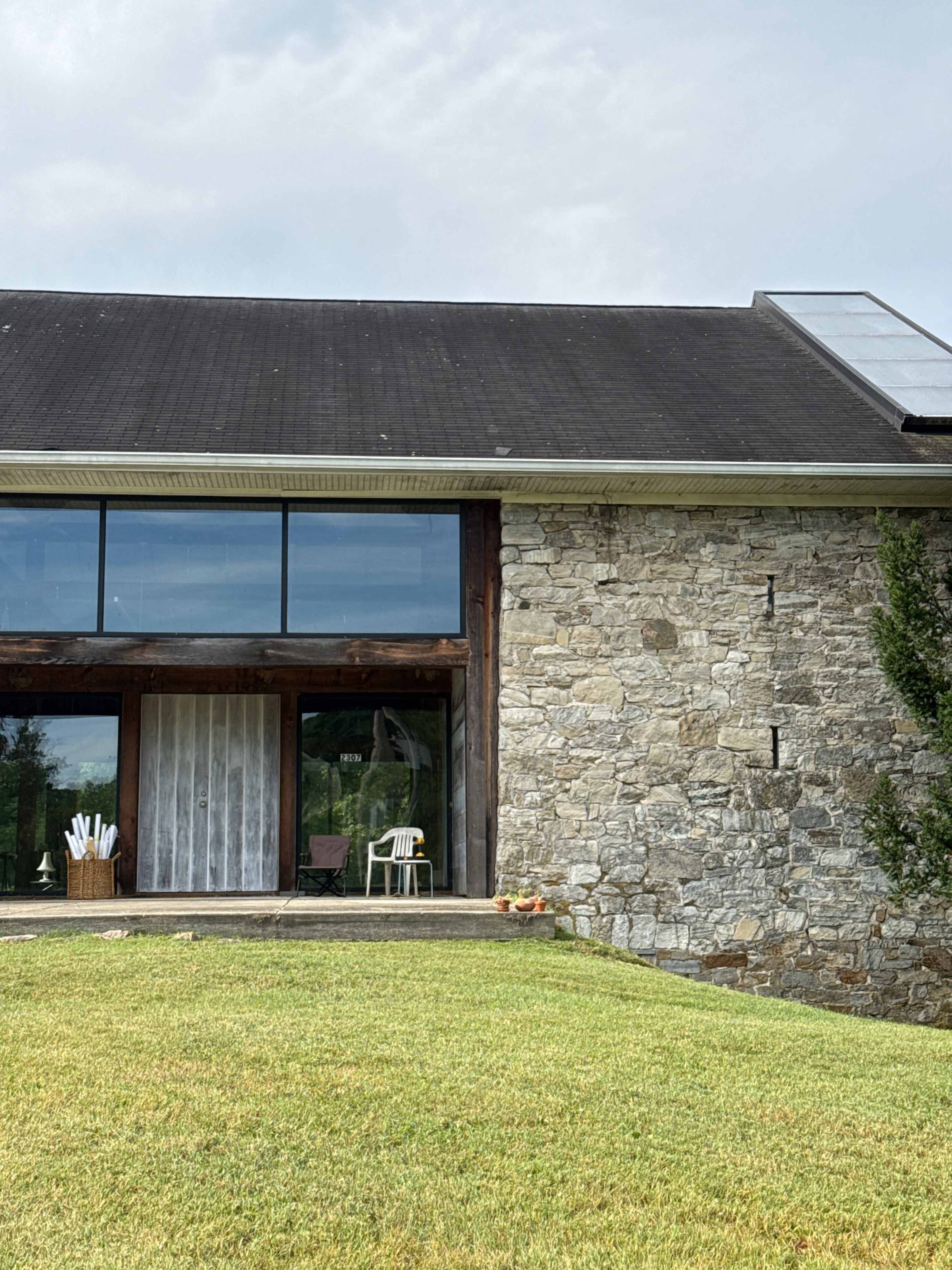 The image shows a modern house with a stone exterior and large glass windows, set against a grassy lawn.