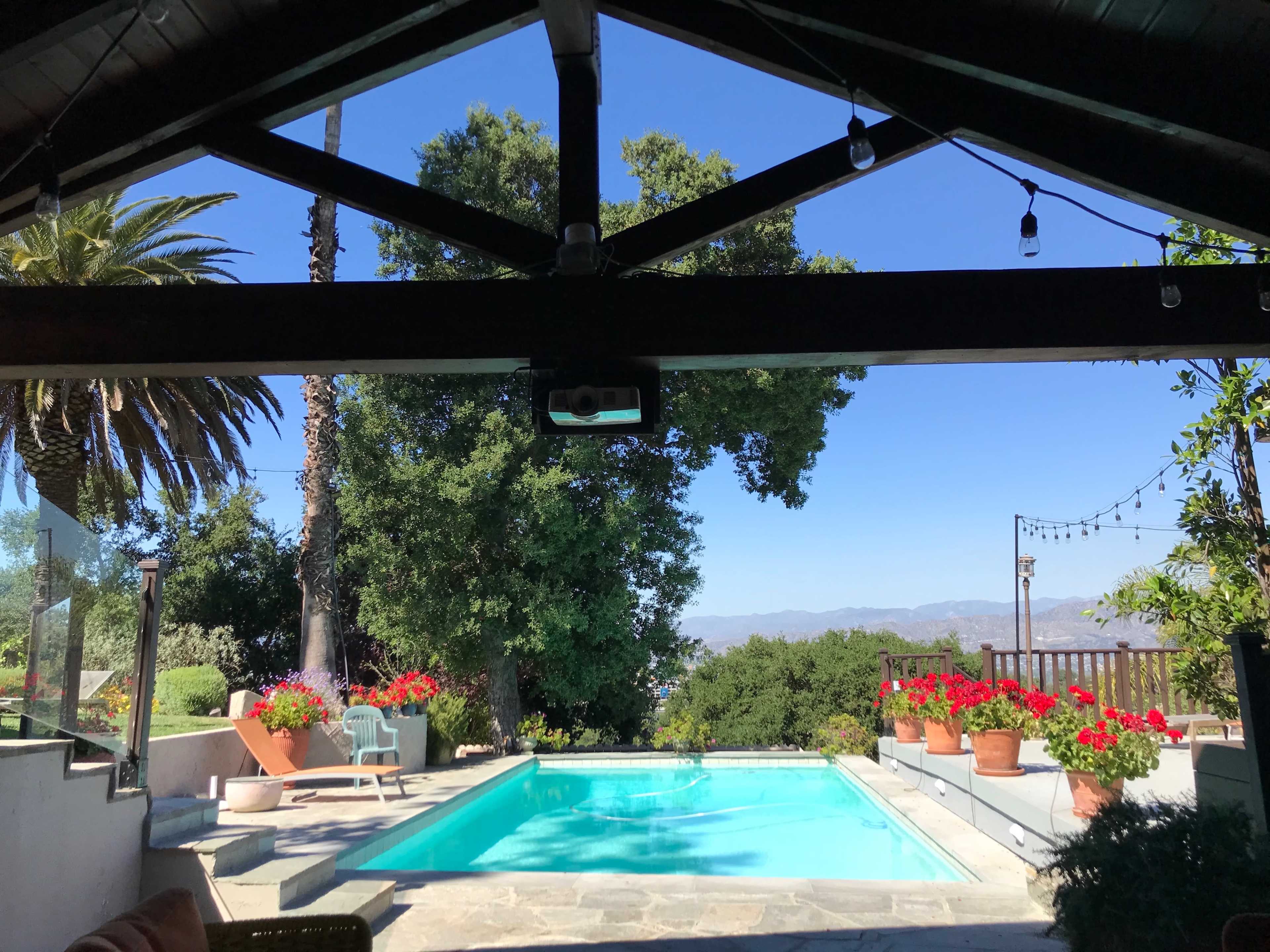 A view from a covered patio looks out over a swimming pool surrounded by potted flowers and trees, with mountains in the background and a clear blue sky.
