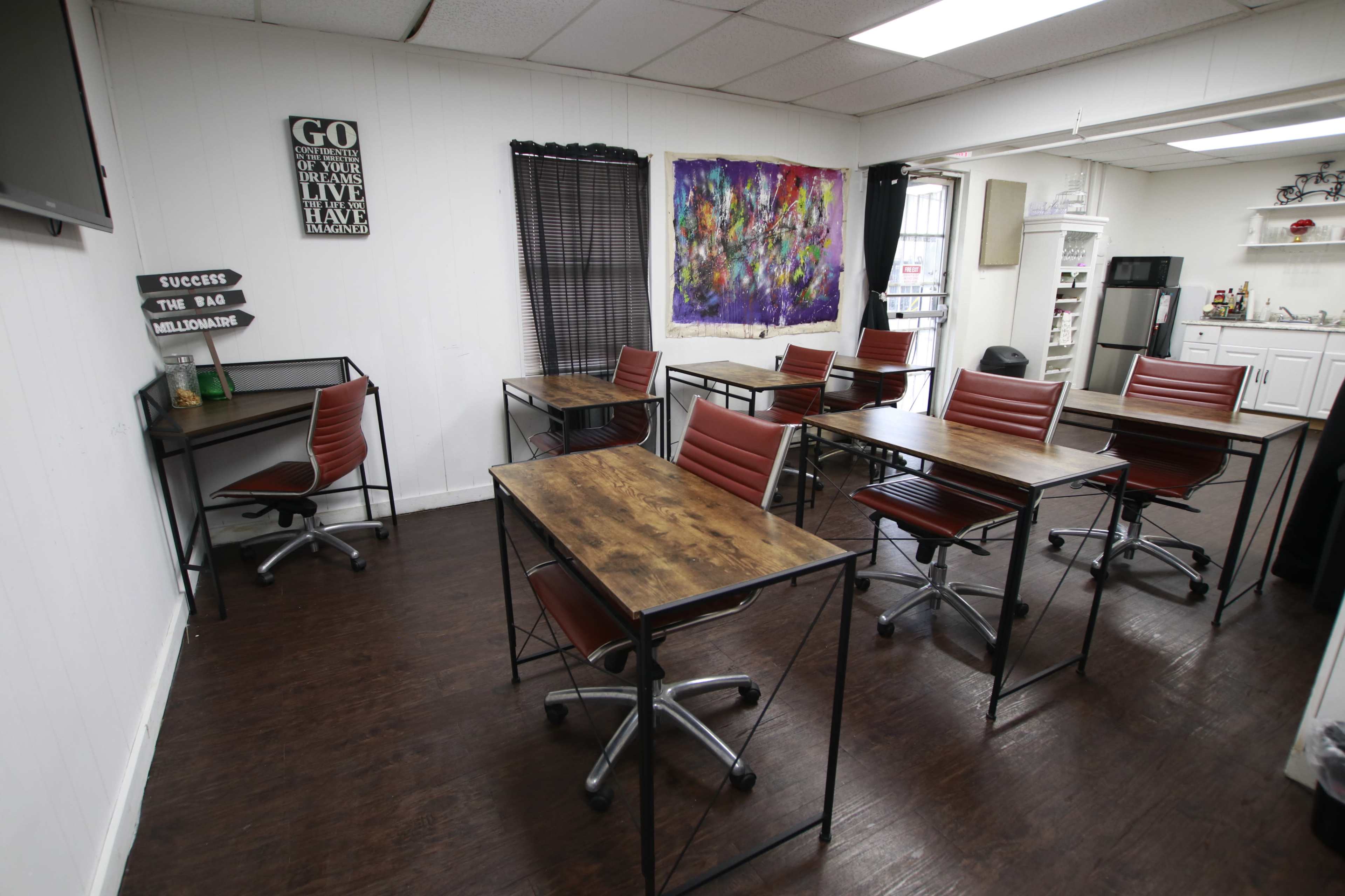 A study or workspace with multiple wooden desks and red chairs arranged in a bright, open room containing a kitchenette area and colorful wall art.